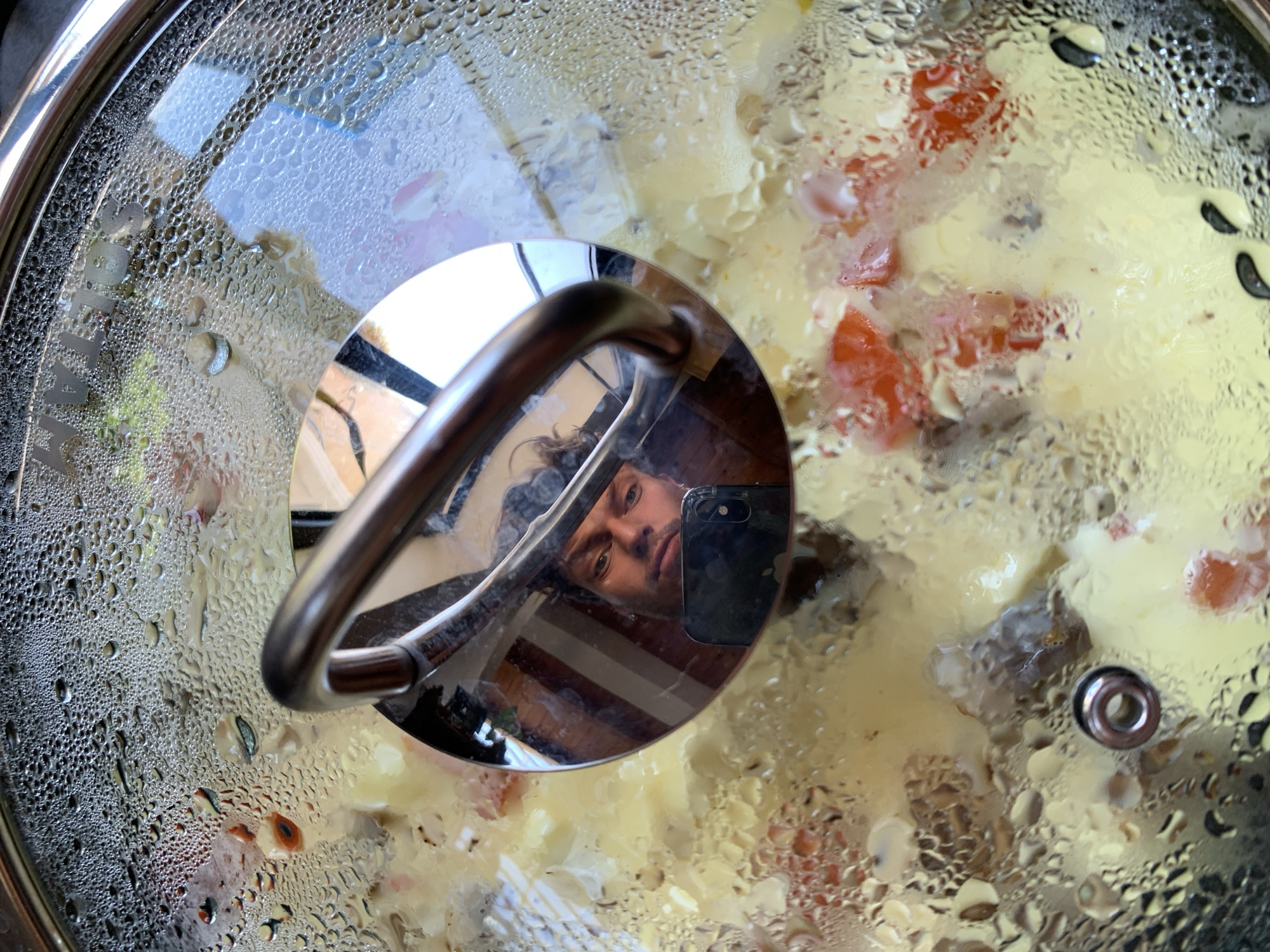 The image shows a pot with a glass lid, presumably on a stovetop, containing food. The lid is covered in condensation. The food inside appears to be a mixture of light-colored, possibly yellow, ingredients with chunks of red and darker elements interspersed. The handle of the lid is reflecting the face of a man holding a phone, suggesting he is taking the photo. There are also reflections of what appears to be a window and possibly striped blinds or wooden panels in the handle. The scene is indoors and the specific location in Tel-Aviv, Israel is not discernible from the provided image.