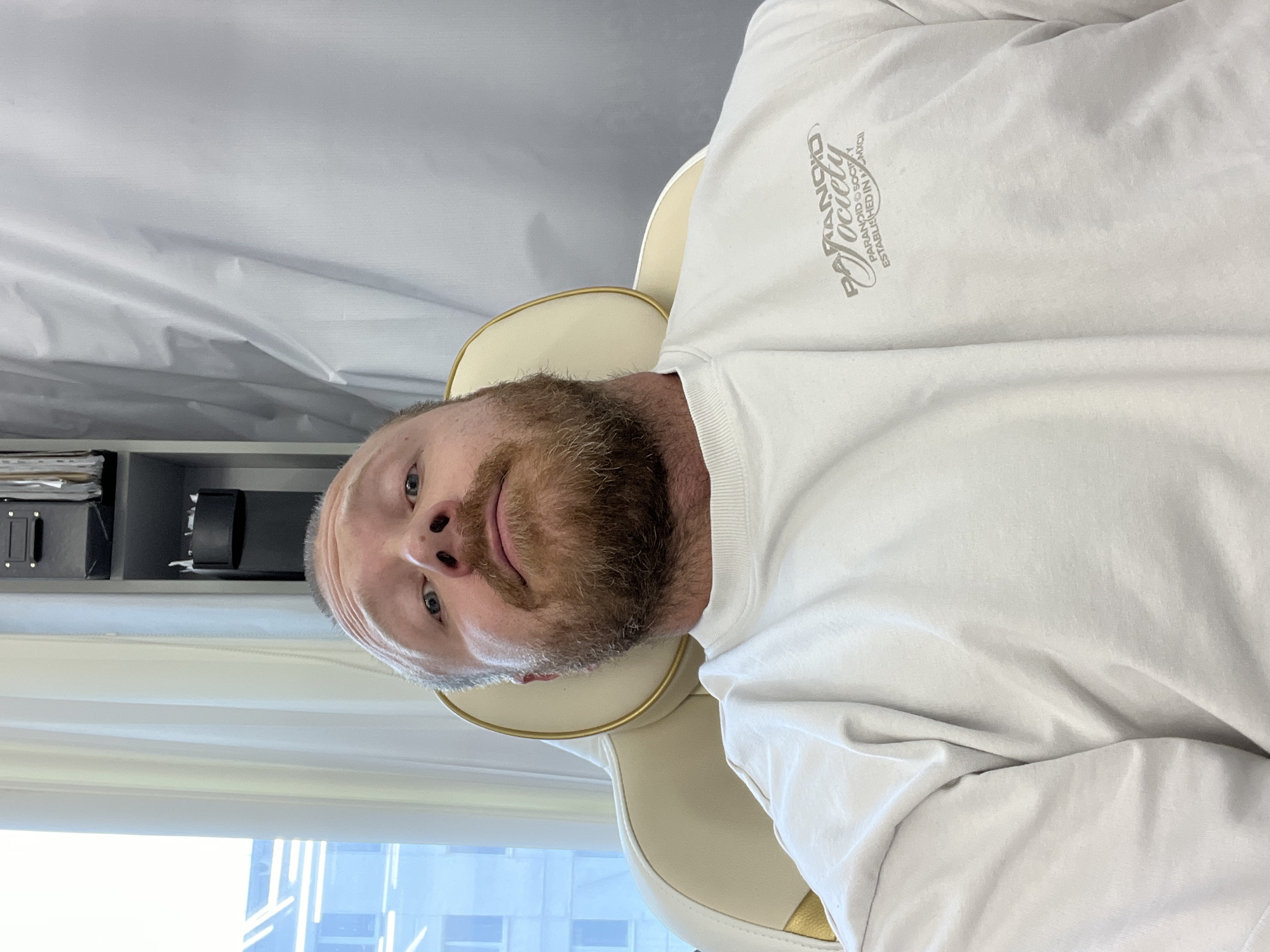 A man with a beard is sitting in an office chair wearing a white long-sleeved shirt with writing on the upper left chest. The office setting includes a shelf with boxes behind him and a window showing a building facade outside. The location is Chaikovsky, Russia.
