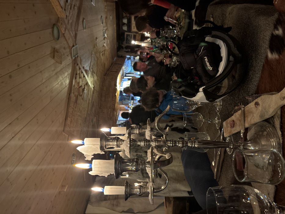 The image captures an indoor dinner scene, likely in the evening, with several people gathered around a long table in what appears to be a rustic restaurant or lodge. The overall lighting is dim and warm, primarily emanating from lit candles.
In the foreground, a prominent silver candelabra with several white candles is centered on the table. Many of the candles have melted wax dripping down, indicating they have been burning for some time. Glasses of various sizes are scattered across the table, some appearing to be wine glasses. A wooden serving board with a slice of cured meat or cheese is visible on the right. The table is covered with dark, furry textiles, possibly animal hides or faux fur, adding to the rustic ambiance.
Several individuals, including adults and at least one child (wearing a blue top), are seated along the table. Most are seen from behind or profile, engaged in conversation or eating. Their postures suggest a relaxed and social atmosphere. On the table, near the people, a black ski helmet with a white rim and clear visor is placed. The brand name "Reusch" is clearly visible in white text on the side of the helmet.
The interior architecture features a ceiling and upper walls clad in light-colored wooden planks, while a lower section of the wall appears to be constructed from exposed brickwork. The style reinforces the alpine or lodge setting. Given the presence of the ski helmet and the provided context of Val-d'Isère, France, the scene strongly suggests a meal following a day of skiing or winter activities in a mountain resort.