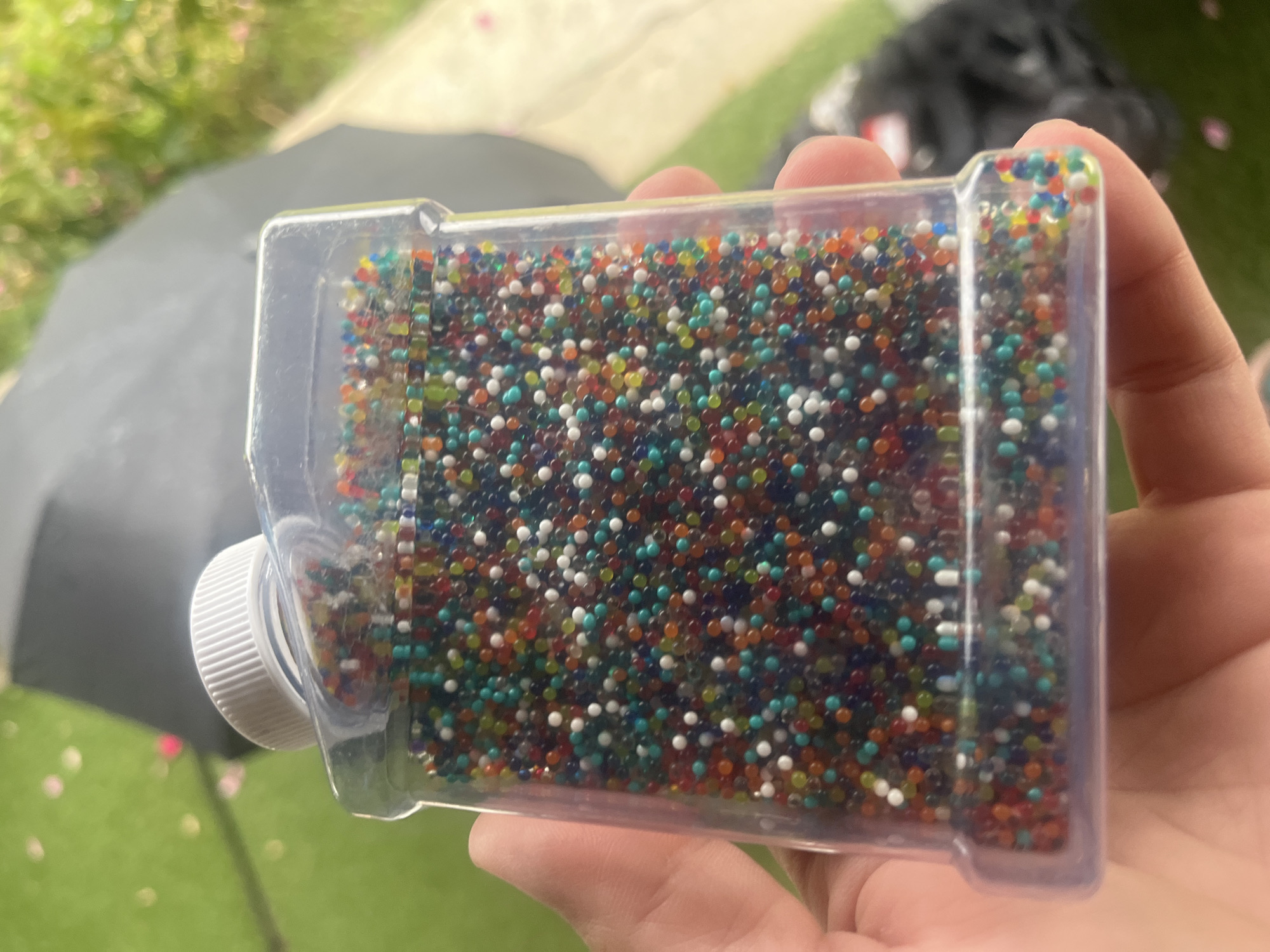 A hand holds a clear plastic container filled with small, colorful beads. The beads are round and vary in color, including red, orange, yellow, green, blue, and white. The container has a white threaded lid. The background is blurred, showing a green, grassy surface, possibly artificial turf, with some pink petals scattered on it. There is also a dark, umbrella-like object and some out-of-focus foliage. The scene is outdoors, and the lighting suggests daytime. The location is not determinable from the image.