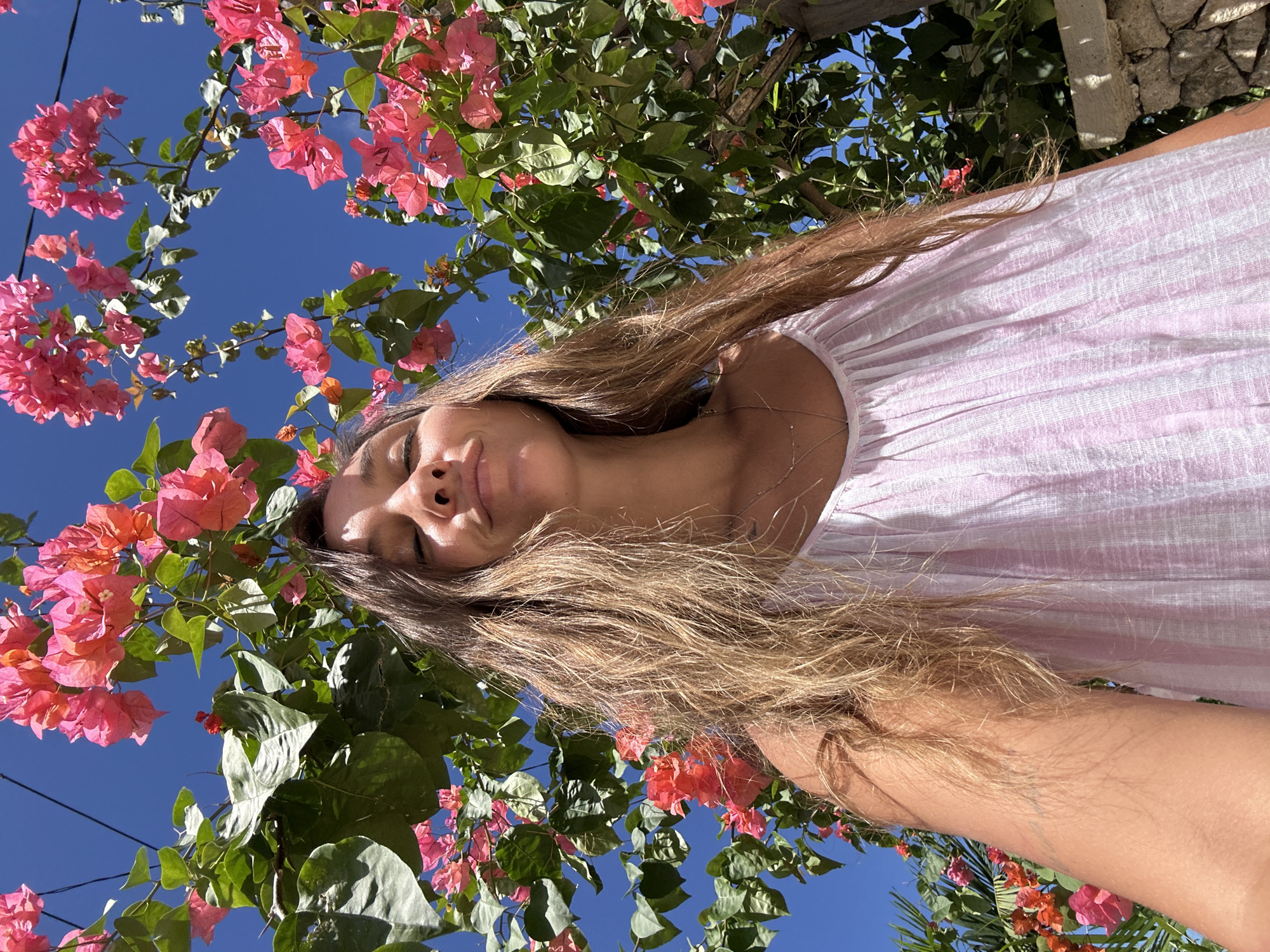 A low-angle shot shows a woman in a pink dress against a backdrop of blue sky and vibrant pink bougainvillea flowers and green foliage. The woman's face is centered, her eyes are closed. Her hair, light brown in color, fans out around her face and chest. The pink bougainvillea surrounds her face. The bright blue sky is visible. The shot may have been taken in Nusa Penida, Indonesia, but the setting cannot be confirmed without additional information.