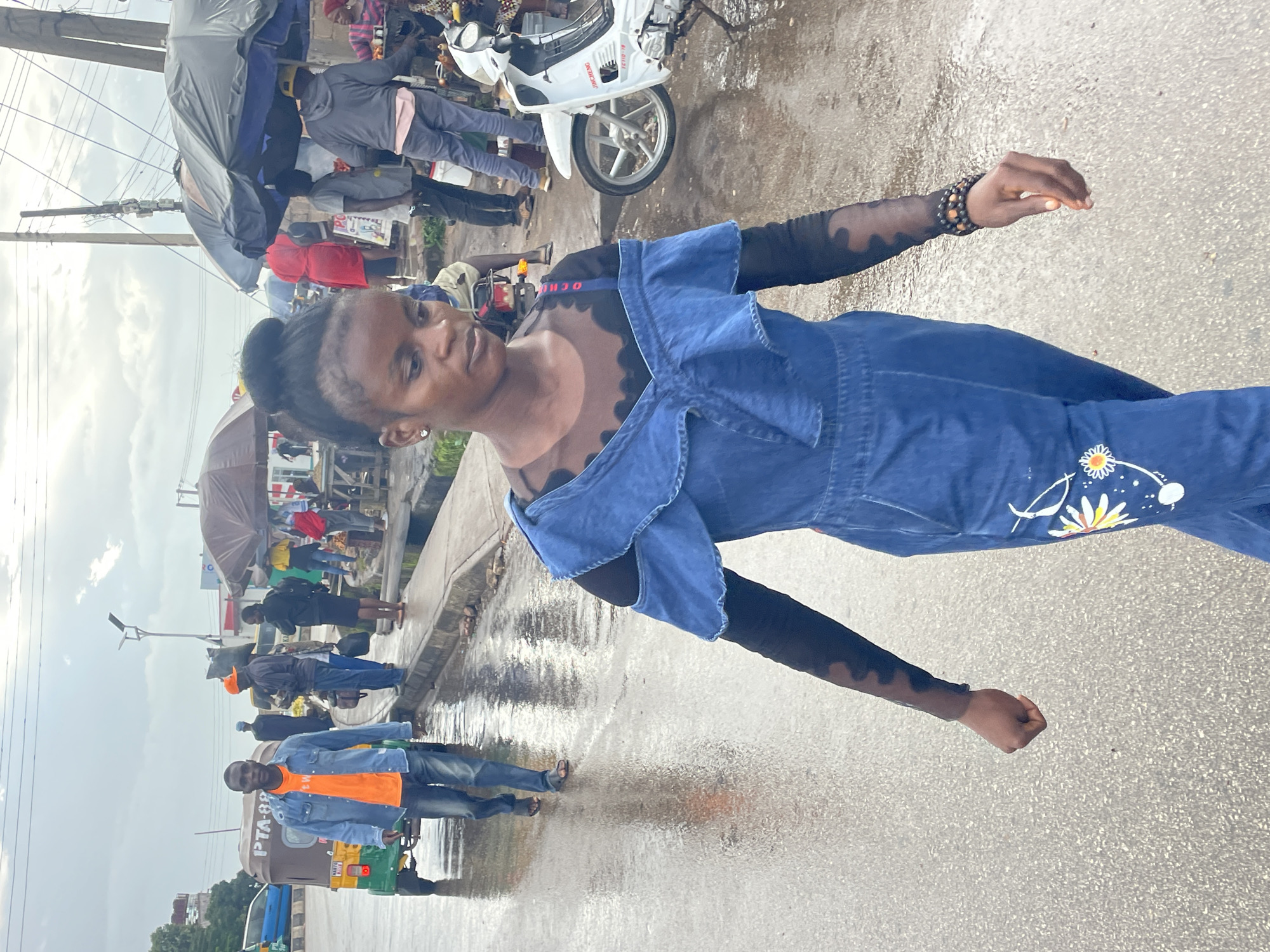 The image depicts a street scene in Jos, Nigeria. The foreground is dominated by a young woman in a blue denim jumpsuit with a black long-sleeved mesh top underneath. She is walking towards the viewer, her left arm bent and hand near her chest. She wears beaded bracelets on her right wrist. The jumpsuit has embroidered floral designs on the leg.
In the mid-ground, a man in a denim shirt and orange t-shirt walks away from the viewer, accompanied by a three-wheeled vehicle. Other individuals and market stalls are visible further back, under umbrellas, with a white motorcycle parked on the right. The ground is wet, indicating recent rain. The sky is overcast. The overall scene suggests a busy, everyday urban environment in Jos, possibly near a market or transportation hub.