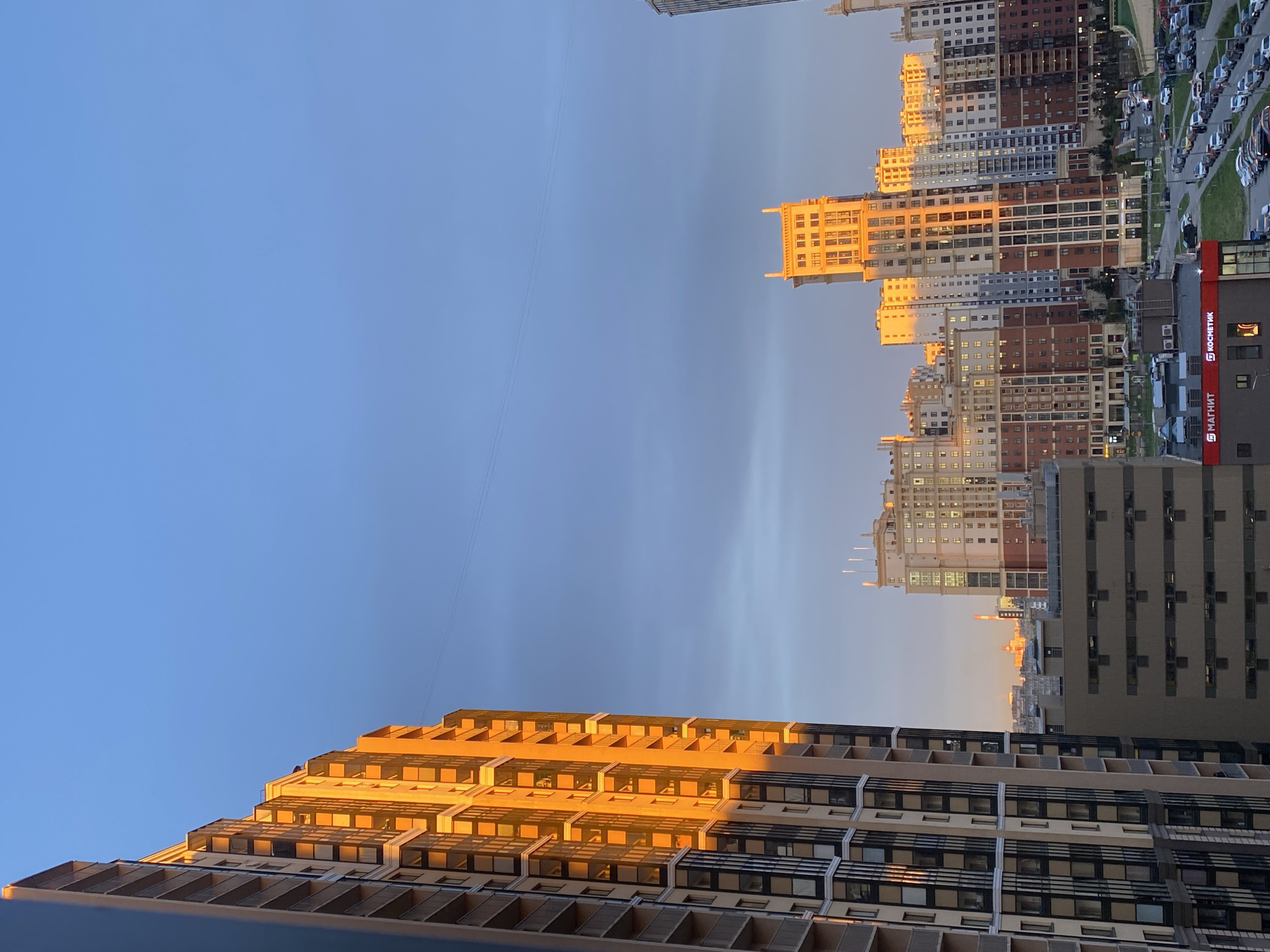 The image shows a cityscape in Saint Petersburg, Russia, taken during the golden hour. The sun's warm light illuminates the facades of several tall residential buildings. One building in the foreground features a tiered design with balconies, prominently lit by the sun. In the background, multiple high-rise apartment buildings create a dense urban landscape. A shopping center with signs in Russian, one of which appears to be "МАГНИТ", is visible. Cars are parked alongside one of the roads. The sky is blue with some thin clouds. An overhead power line is visible against the sky.