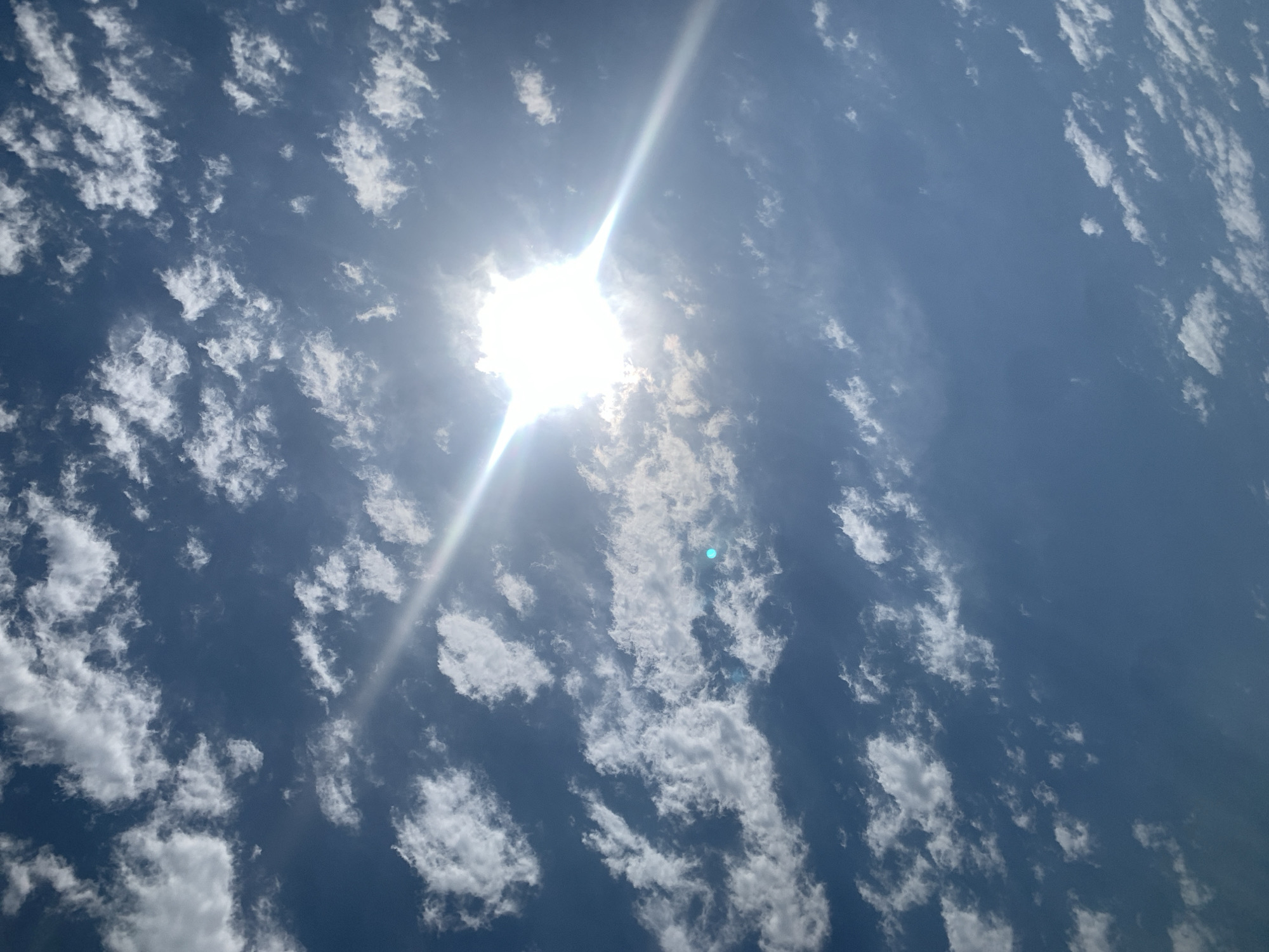 A bright sun, partially obscured by thin clouds, shines down on a blue sky. The sun is a dazzling white disc, with rays extending outwards, creating a lens flare effect. Scattered cumulus clouds, white and fluffy, drift across the sky. The scene is an outdoor shot, likely taken during the day. There are no people or man-made objects visible, focusing solely on the natural elements of the sky and sun. The image captures a moment of clear weather with scattered cloud cover. The specific location of Khazar Raion, Azerbaijan is not visually evident in the image.