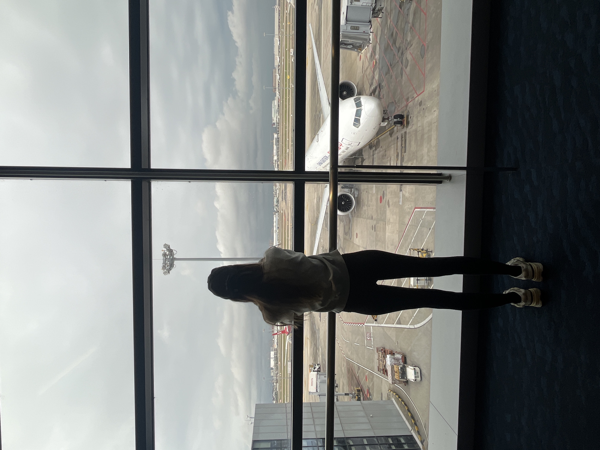 A full-body shot from behind shows a young woman with long, dark hair standing at a large window overlooking an airport tarmac. She is wearing a light-colored long-sleeved shirt, black leggings, and white sneakers. The woman is facing away from the camera, looking out at an airplane parked at a gate, which is connected to a jet bridge. The plane is white with "CHINA EASTERN" written on its side in red lettering. There are also several ground vehicles and other airplanes visible on the tarmac. The sky is overcast and cloudy. The image appears to be taken at Pudong International Airport in Shanghai, China, given the presence of China Eastern Airlines and the typical architecture of a modern international airport terminal.