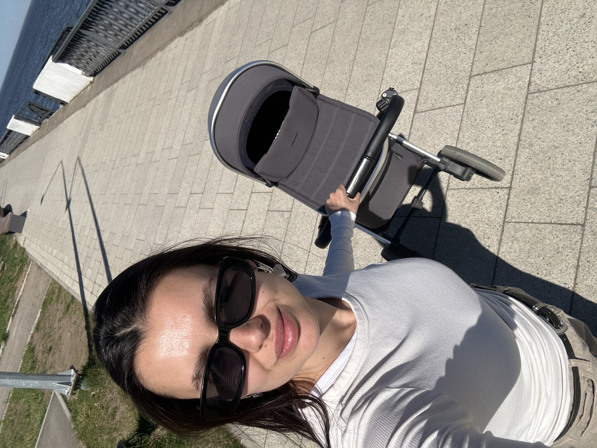 A selfie taken from above shows a woman standing on a light grey tiled walkway next to a river or large body of water. The woman has dark hair and is wearing large black sunglasses and a light-colored long-sleeved shirt. She is holding the handle of a grey baby stroller in front of her. The walkway is bordered by a metal railing and some grassy areas. This image was taken in the city of Chaikovsky, Russia.