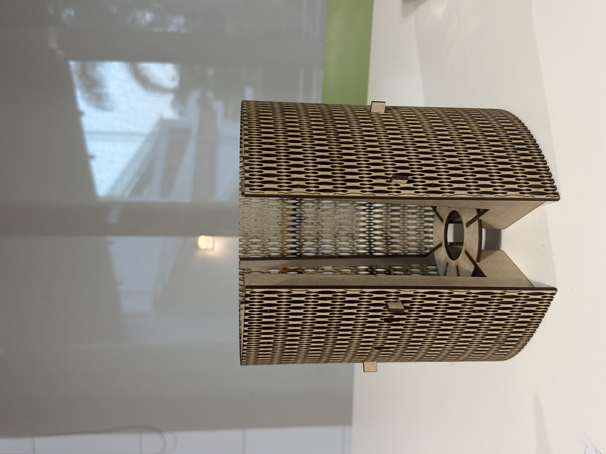 A laser-cut wooden lampshade or decorative light fixture is displayed lying horizontally on a reflective, white surface. The object is crafted from light-colored wood, likely plywood or MDF, with dark, laser-burnt edges outlining an intricate, repeating pattern of elongated, interconnected oval cutouts that form the curved sides. Visible interlocking tabs suggest the piece is assembled from multiple flat components. One end of the fixture shows a central circular opening, presumably for a light fitting.
The white surface beneath the object clearly reflects its form. The background consists primarily of a plain, light-colored wall, with a distinct vertical strip of light green visible on the right side of the frame. Reflections on the wall, possibly originating from a window, reveal blurred and indistinct shapes that could be outdoor scenery, structures, or even a very faint, ambiguous figure or face in the upper left section. The scene is brightly lit, typical of an indoor setting, with ambient light contributing to the reflections. No specific activity is taking place, and no text is visible. The location is San Bernardino Tlaxcalancingo, Mexico.
