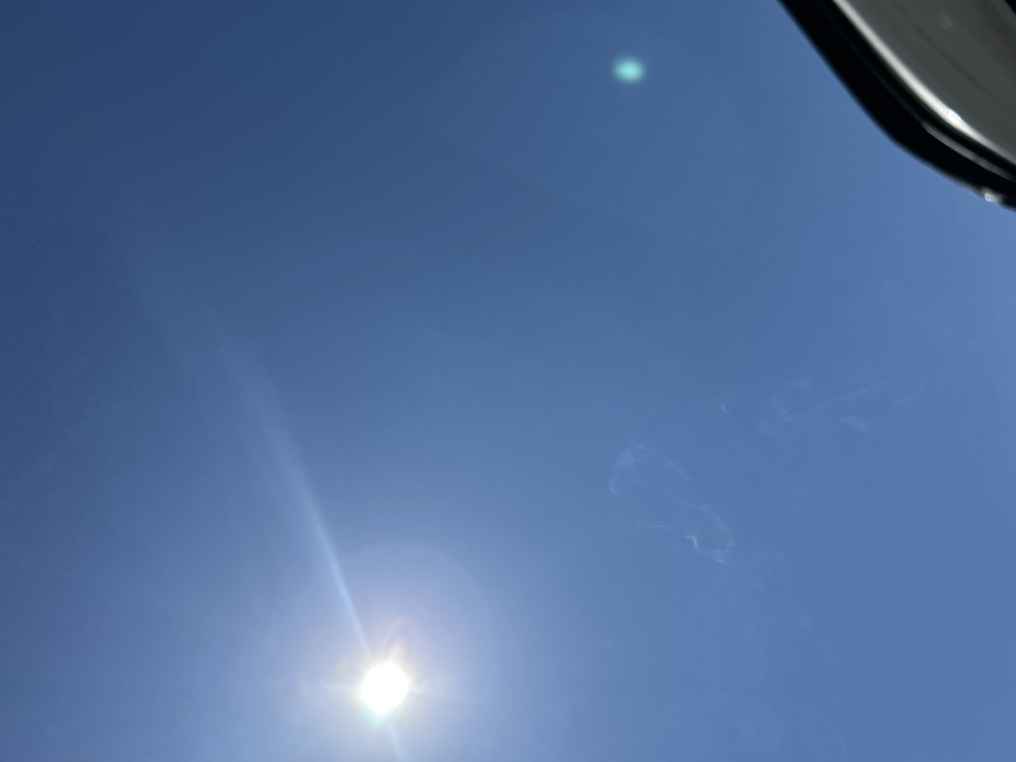 The image depicts a view of a clear, bright blue sky with the sun shining brightly at the bottom left corner. Faint wisps or trails appear to be present in the upper right section of the frame, possibly thin cirrus clouds or contrails. The top right corner shows a portion of an object, potentially a car frame or another vehicle component. It is impossible to confirm that this image was taken in the city of Mineralni Bani, Bulgaria, without additional information.