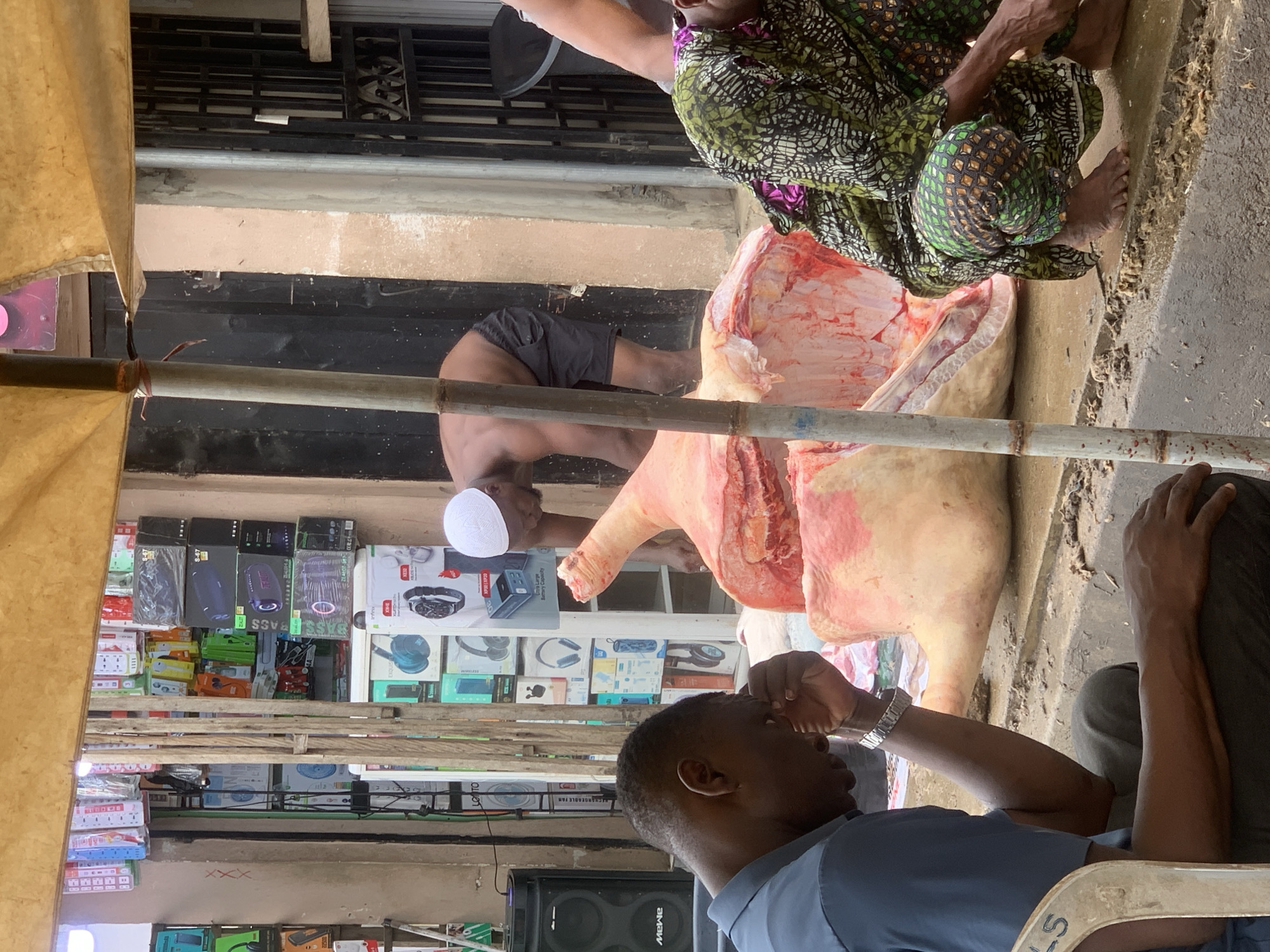 The image depicts an outdoor scene in Ikorodu, Nigeria, centered around the butchering of a large animal carcass, likely a cow. The carcass is partially butchered, revealing raw meat and bone. Three men are present. One man, wearing a white cap and dark shorts, is bent over the carcass, presumably butchering it. Another man is seated nearby in traditional African clothing. A third man, wearing a blue shirt and a wristwatch, is seated in the foreground. Behind the butchering area, a store is visible, displaying electronics in boxes. A yellow tarp is visible, supported by bamboo poles, likely providing shade. The ground appears to be concrete.