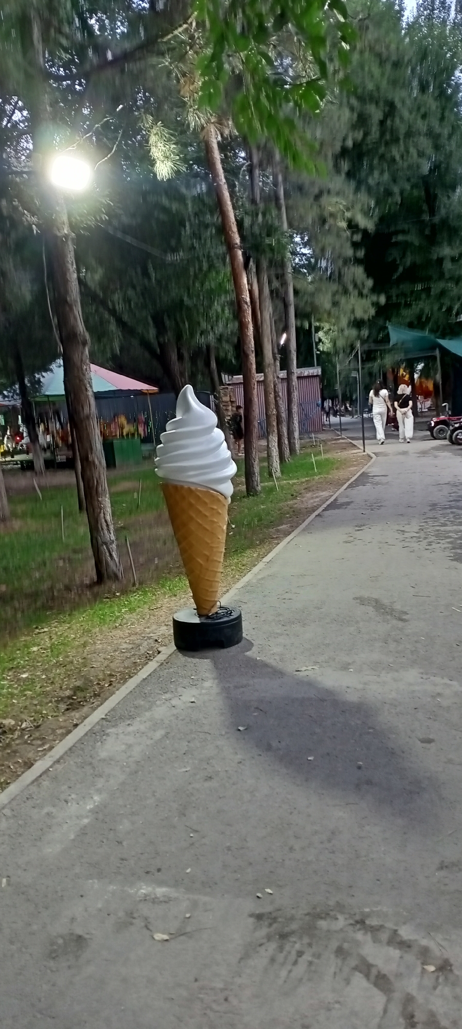 A large, white soft-serve ice cream cone sculpture stands prominently on a paved walkway in what appears to be a park or recreational area. The sculpture is supported by a black, circular base, possibly a tire. The walkway is lined with trees, some of which are illuminated by a bright overhead light. In the background, two women in white clothing are walking away from the camera, and other indistinct figures and structures, including what looks like a carousel, are visible further down the path. The environment is green with grass and trees, suggesting an outdoor setting in Sokuluk, Kyrgyzstan. The overall lighting suggests it is either dusk or an overcast day.