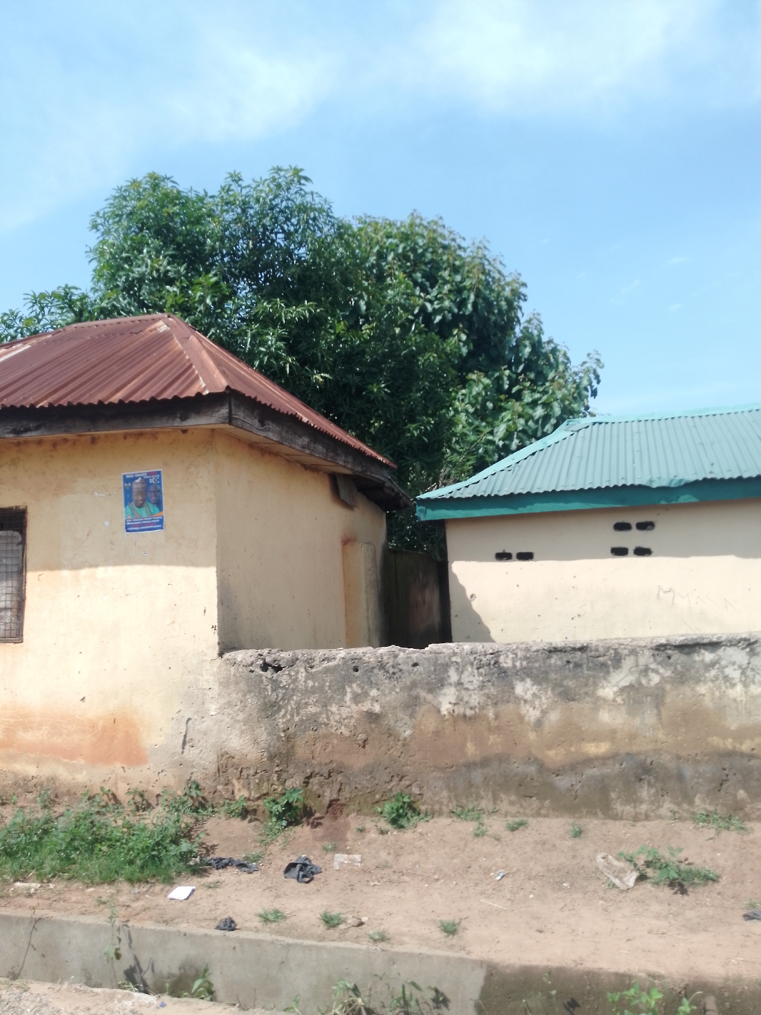 The image shows two buildings with corrugated metal roofs, one rusted brown and the other a pale green, under a clear blue sky. The walls of the buildings are a light yellow, and one has a poster on it. In front of the buildings is a low, rough concrete wall. Beyond the wall, the ground is dirt and sparsely covered with dry grass and weeds. There is also some debris visible on the ground, including a few pieces of black cloth and a white piece of paper. The scene appears to be in a rural or semi-urban area of Serti, Nigeria, characterized by simple construction and a dry environment.