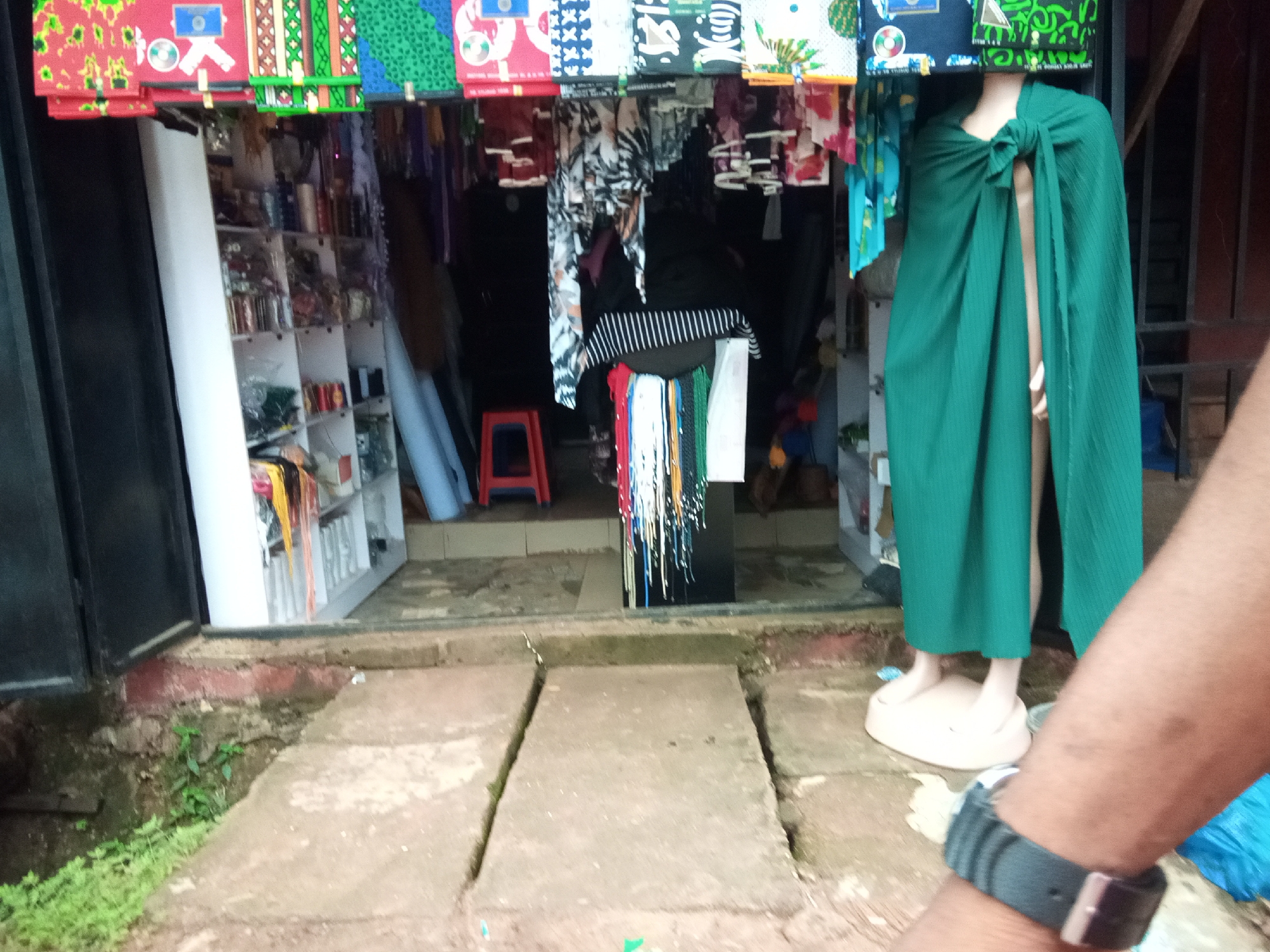 The image shows a street scene in Oko Town, Nigeria, with a focus on a small shop entrance. In the foreground, a person's arm with a watch on the wrist is visible, indicating a first-person perspective. The shop displays various fabrics and textiles, hanging from the ceiling and displayed on shelves. A mannequin is positioned outside the shop, draped in a green fabric. The ground in front of the shop is paved with concrete slabs. The overall environment suggests a marketplace or commercial area, with a mix of indoor and outdoor elements.
