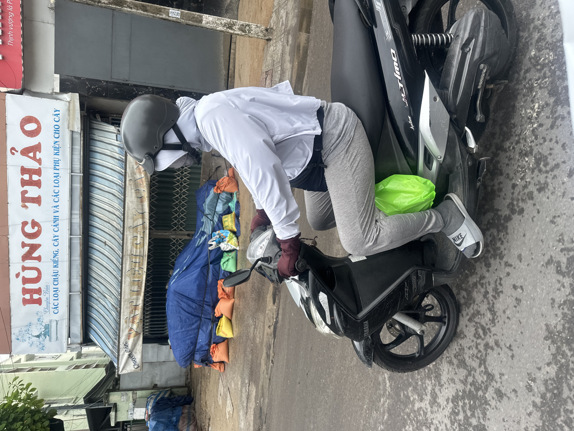 The photo shows a person wearing a helmet, white jacket, gray pants, and Nike slides sitting on a Yamaha scooter. The person is leaning forward, as if about to start the scooter. A green plastic bag is placed between the person's legs and the scooter. The scene appears to be set in the city of Bac Nha Trang Ward, Vietnam, as suggested by the signs in the background, which are in Vietnamese. One sign reads "HÙNG THẢO" and includes the text "CÁC LOẠI CHẬU KIỂNG, CÂY CẢNH VÀ CÁC LOẠI PHỤ KIỆN CHO CÂY," indicating it's a business specializing in pots, ornamental plants, and accessories for plants. Behind the person and the scooter, there is a closed storefront with a striped awning, with stacked bags and supplies covered with a blue tarp. The street and sidewalk appear to be made of concrete.