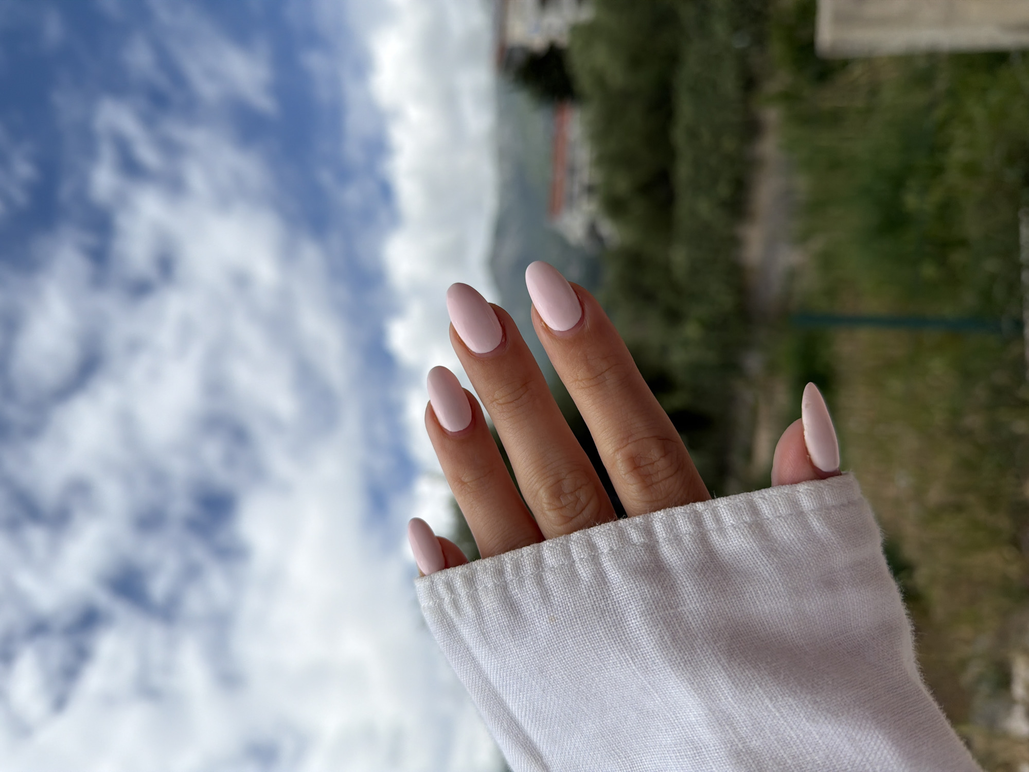 A hand with light pink, almond-shaped nails is positioned against a backdrop of a cloudy sky and blurred greenery. The hand is partially covered by a white, linen-like sleeve. In the background, indistinct building structures can be discerned amidst the foliage. Given the landscape featuring a blend of mountainous terrain and coastal vegetation, combined with building styles present, the location is possibly Budva, Montenegro.