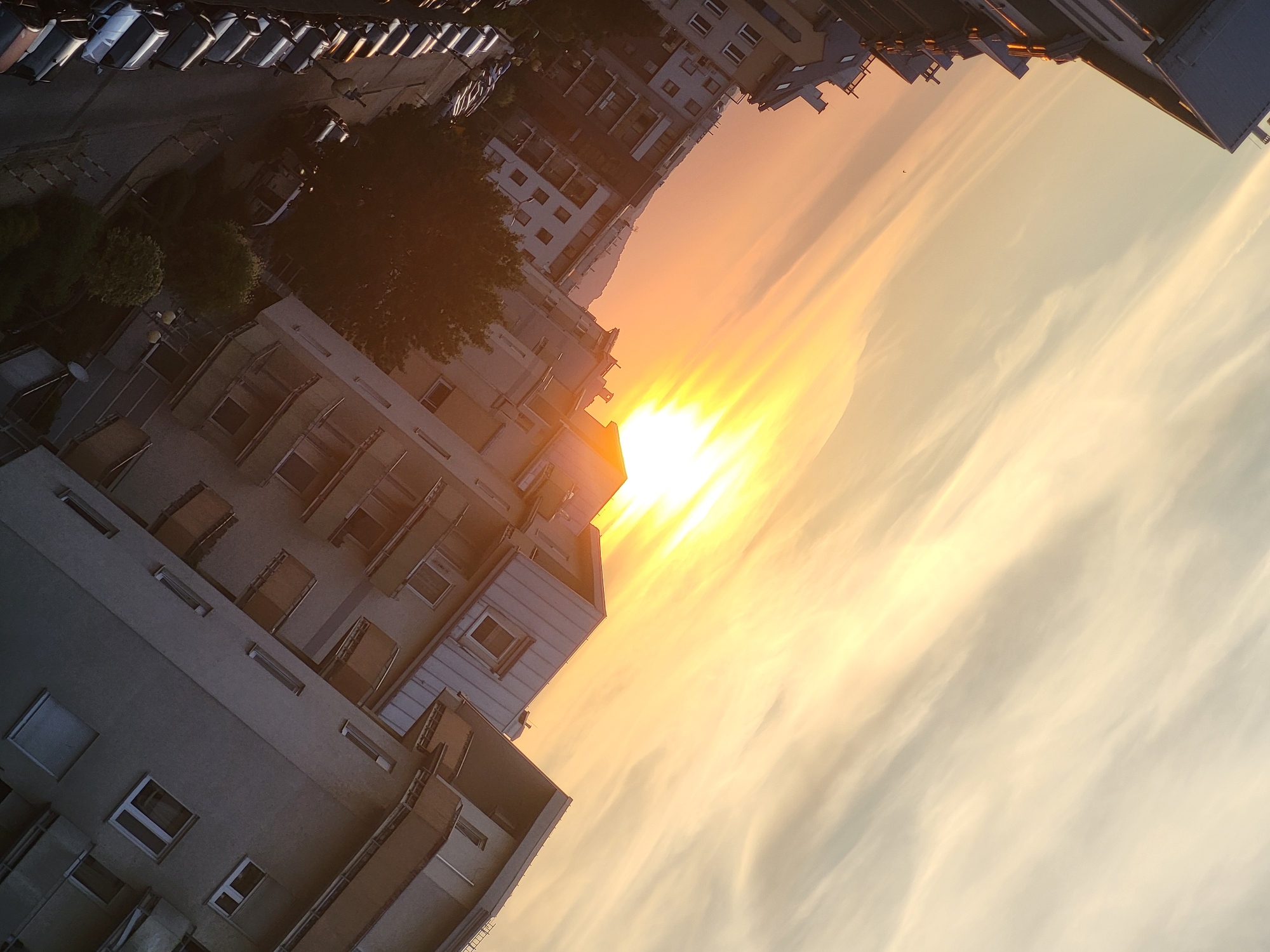 The image depicts an urban landscape at sunset, with buildings and parked cars dominating the foreground and middle ground. The sun is low in the sky, casting a warm, golden glow across the scene and creating long shadows. The sky is filled with streaks of orange, yellow, and white clouds, indicating the setting sun. The buildings are multi-story structures with visible windows and balconies. A street lined with parked cars is visible in the lower left portion of the image. The perspective is tilted, giving a dynamic feel to the composition. The overall impression is one of an urban evening, bathed in the soft light of sunset.