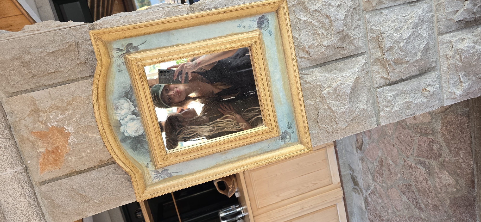 A framed mirror hangs on a stone wall with pale-colored stone blocks and a section with red stone. The frame of the mirror is gold with blue floral decorations. The mirror reflects a person holding a phone taking a selfie and another person below them. In the background, a wooden cabinet is visible. The location is Bogišići, Montenegro.