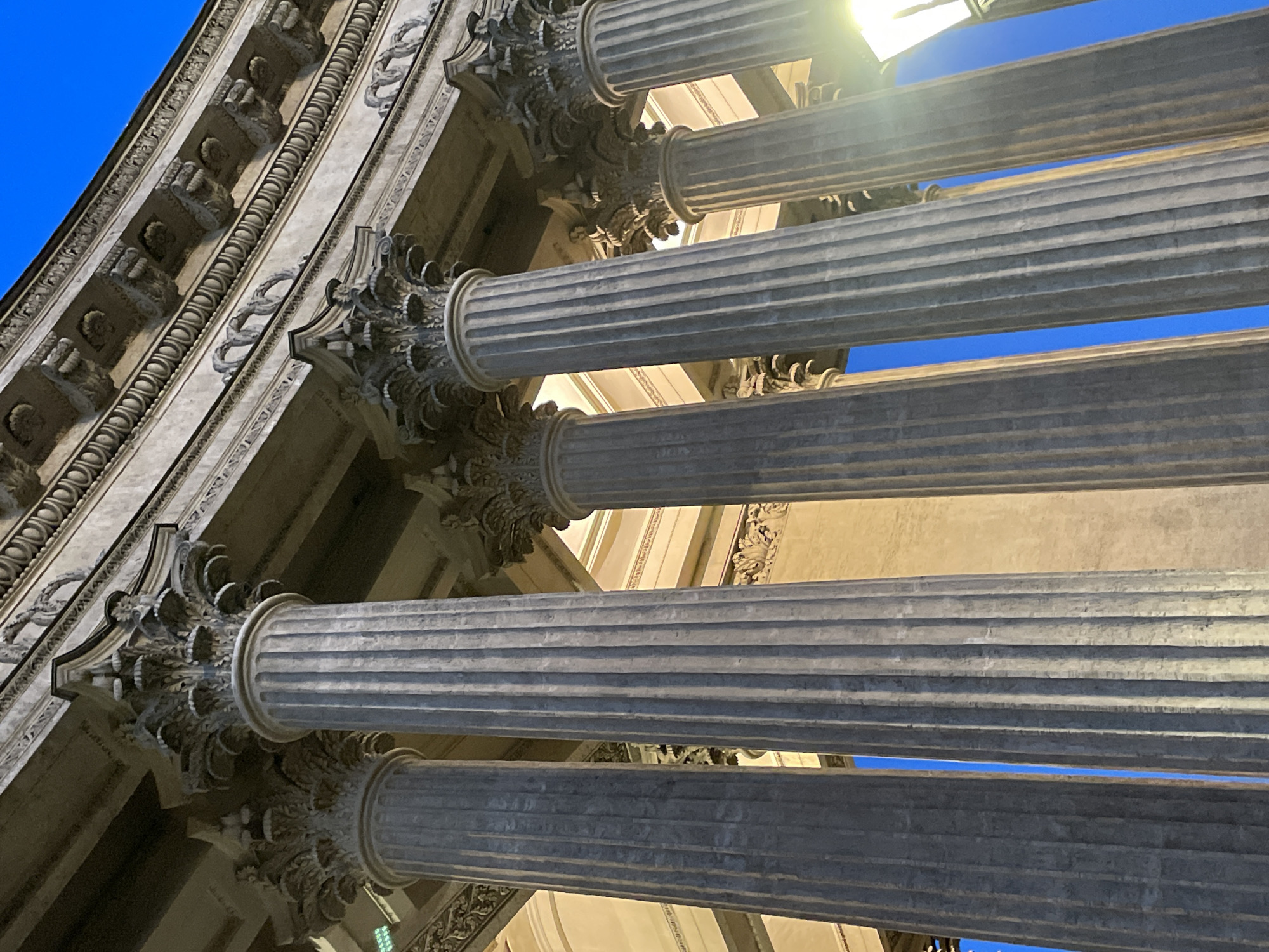 The image captures a low-angle, upward view of a grand classical building, likely located in Saint Petersburg, Russia. The primary subjects are several towering, fluted columns adorned with elaborate Corinthian capitals, featuring intricate acanthus leaf designs. Above the capitals, a richly decorated entablature is visible, showcasing multiple bands of ornate carvings, including corbels, egg-and-dart patterns, and looping motifs.
The architectural elements appear to be constructed from light-colored stone, possibly marble or granite, exhibiting detailed texture and some signs of weathering. A vibrant, clear blue sky serves as the background, visible through and above the columns, indicating a bright, sunny daytime setting. A prominent lens flare from the sun is visible in the upper right portion of the image, illuminating parts of the columns and the sky. The interior of the structure, visible beyond the columns, appears to be in shadow, contrasting with the brightly lit exterior. No people or visible text are present in the frame. The photograph highlights the impressive scale, intricate detailing, and classical aesthetics of the architecture.