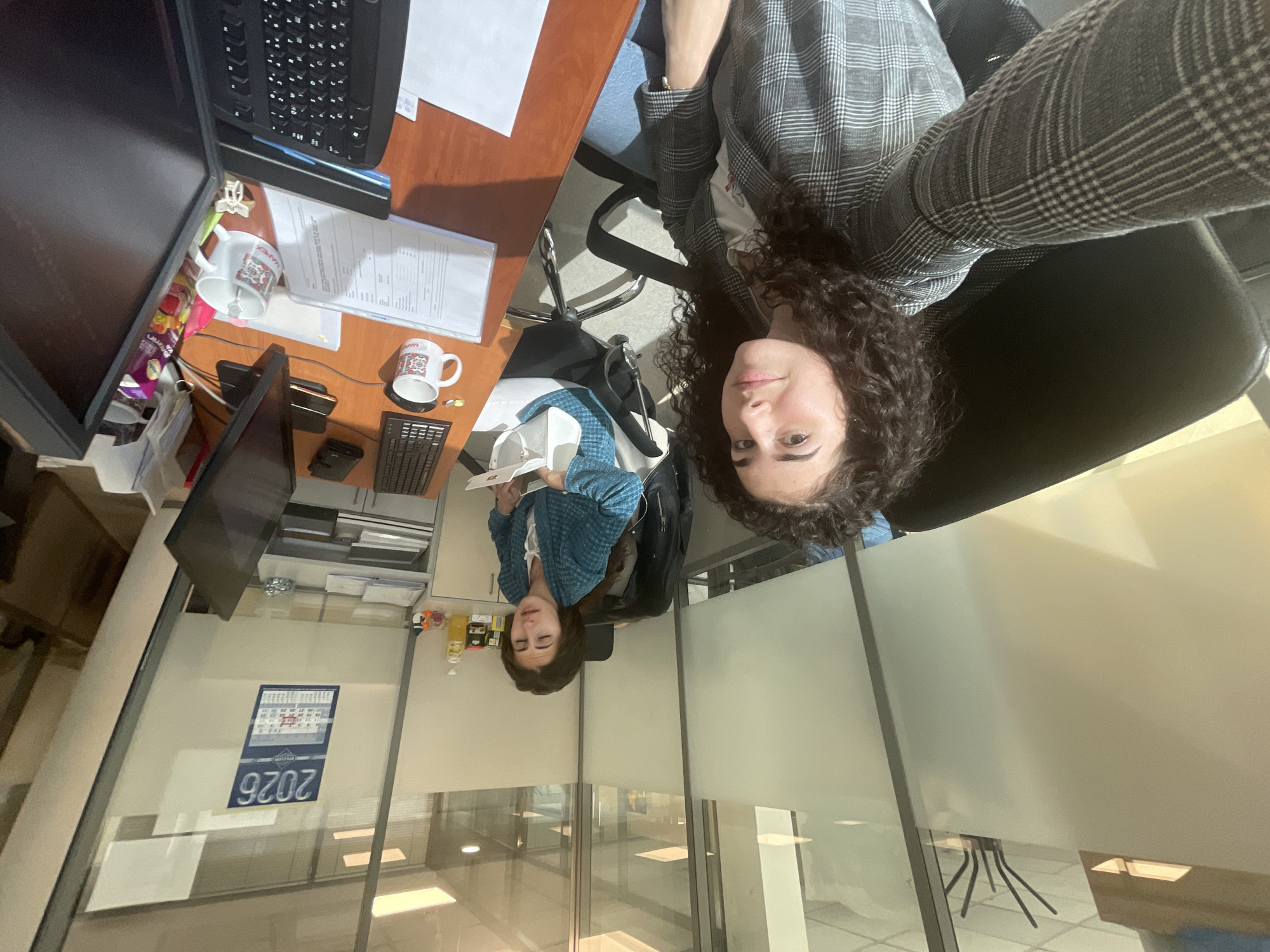 A person with curly hair is taking a selfie in an office environment. The office has cubicles with frosted glass partitions. A calendar visible on a partition indicates the year 2026. Office supplies, including a computer, keyboard, monitor, mugs, and papers, are visible on a desk. The person is wearing a blazer and appears to be sitting in an office chair. Based on the "АВУЕРИНС" text on the calendar, the location might be Sofia, Bulgaria.