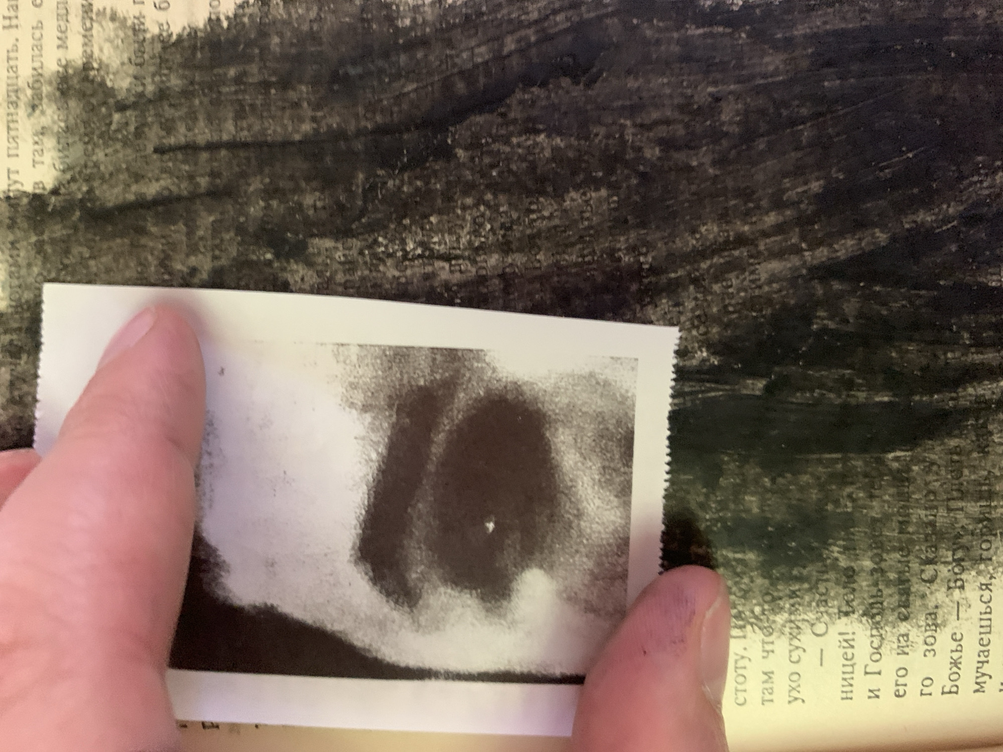The image shows a close-up of a hand holding a black and white photograph over a textured, dark background that appears to be a page from a book or a canvas with brushstrokes. The photograph, partially obscured by the hand, is a close-up of what looks like an eye or a dark, amorphous shape with a bright speck in the center. The background is a dark, abstract pattern, possibly paint or ink, with some lighter areas that suggest texture or perhaps faint writing.
The text visible on the book page, in Russian, mentions "пятнадцать" (fifteen), "счастье" (happiness), "Господь" (Lord), and "Божье" (God's). The context of the text suggests a narrative or reflective content, possibly religious or philosophical. The location context provided is Ganino, Russia. The overall mood is introspective, with the combination of the personal photograph, the textured background, and the evocative text.
