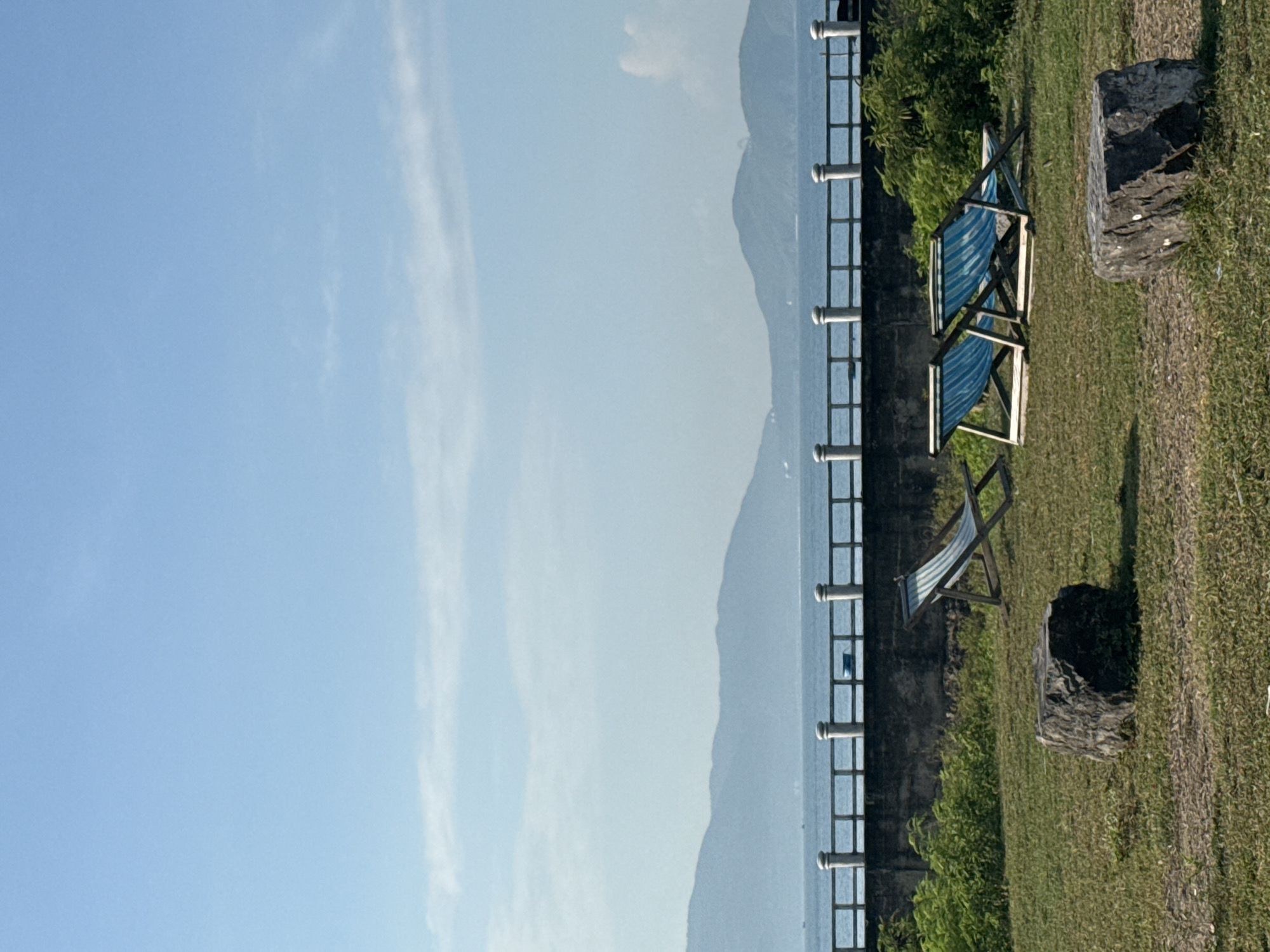 This is a vertical, eye-level, outdoor photograph of a serene waterside landscape. In the foreground, two large, dark, irregular rocks sit on a patch of dry, brown grass. Behind the rocks, a section of manicured green grass slopes gently upward. Two blue and white striped deck chairs are positioned on the grass; one is angled to the left, and the other two are side-by-side facing right. Behind the chairs is a dense patch of vibrant green bushes.
A low, dark concrete wall runs horizontally across the mid-ground of the image, separating the grassy area from the water. A white railing with evenly spaced posts runs along the top of the wall. Beyond the wall, a calm, blue body of water stretches towards the horizon.
In the distance, a range of hazy blue mountains forms the horizon line under a clear, pale blue sky with wisps of white clouds. The overall impression is one of tranquility and natural beauty. While the image does not definitively place the scene in Bac Nha Trang Ward, Vietnam, the tropical vegetation, mountains, and coastal setting are consistent with many locations in Vietnam.