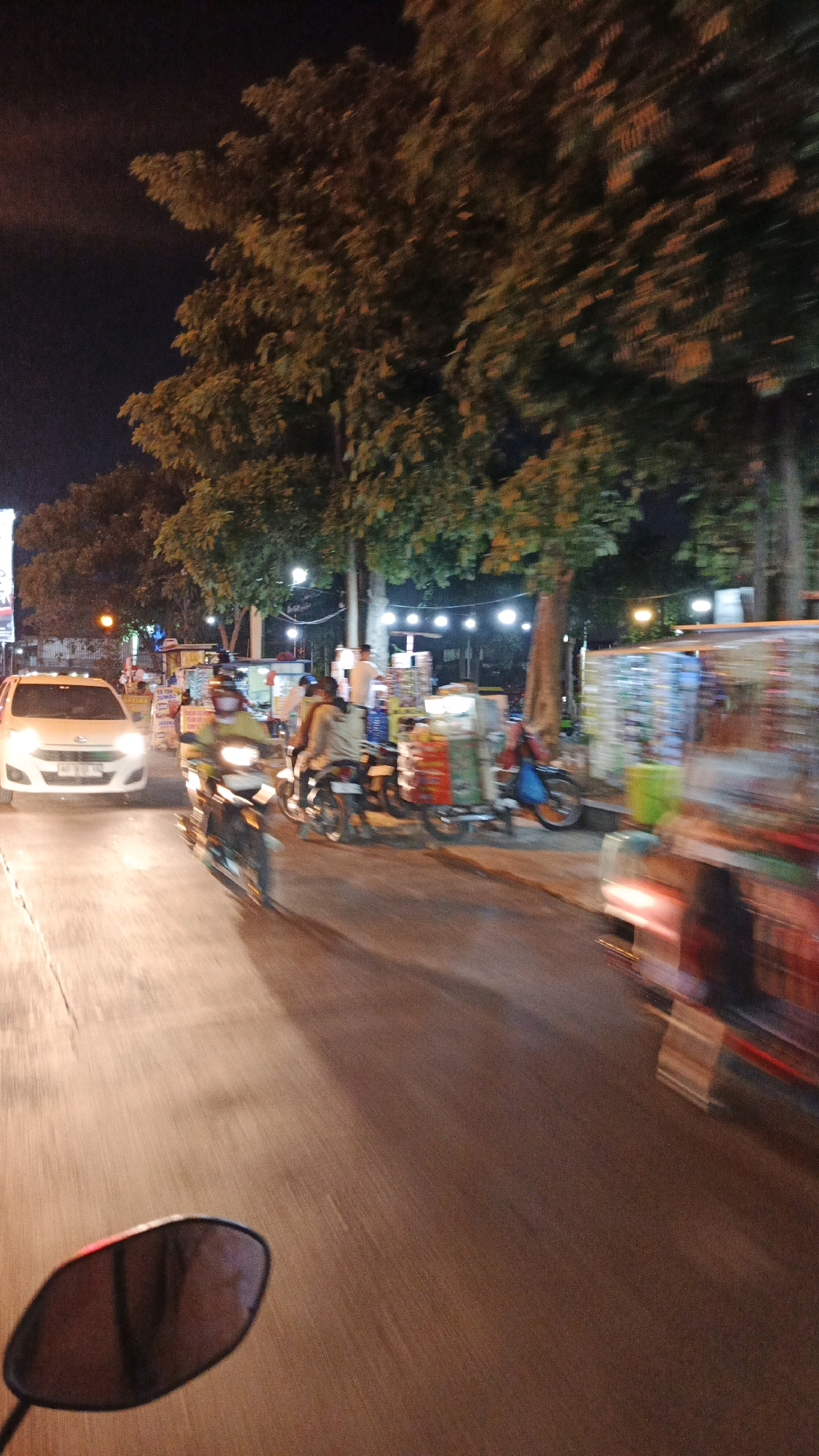 The image depicts a street scene at night in Surakarta, Indonesia, characterized by motion blur suggesting a moving vehicle, likely a motorcycle from which the photo was taken, given the presence of a rearview mirror in the lower left. A white car with its headlights on is visible on the left side of the road. Several motorcycles are also present, with riders, and some appear to be moving. On the right side of the road, numerous stalls with colorful displays, possibly vendors, are set up along the sidewalk, illuminated by string lights overhead. Trees line the street, their foliage also blurred by the motion. The overall impression is a lively, bustling night market or street food area.