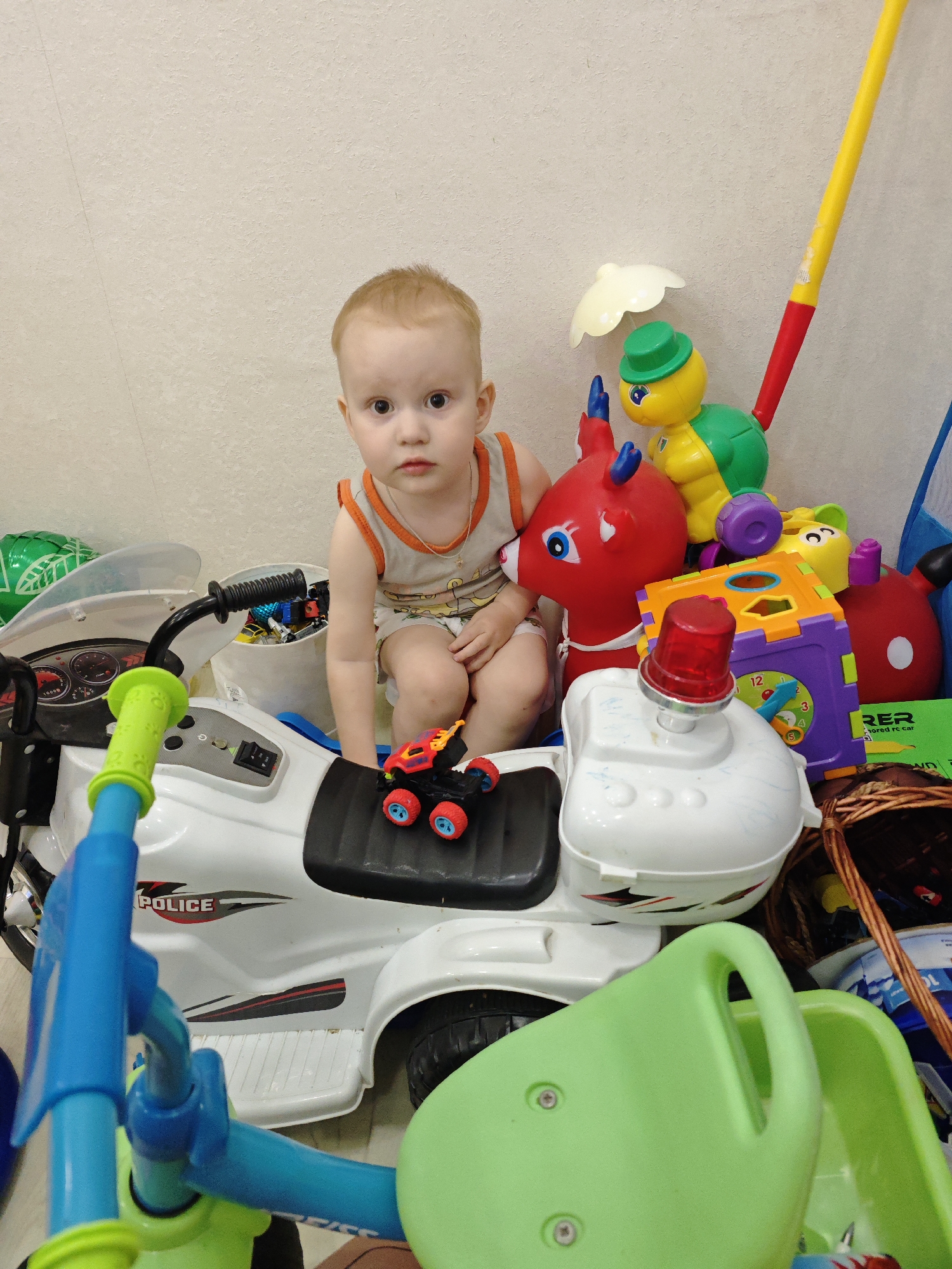 A young child with fair skin and reddish-blond hair is seated atop a white toy police motorcycle, surrounded by various toys. The child wears a light-colored sleeveless shirt with orange trim and matching shorts. The motorcycle has black accents and "POLICE" printed on its side. A small toy car rests on the motorcycle's seat.
Toys include a red inflatable deer, a yellow and green turtle pull-along toy, and a multicolored shape sorter cube with a clock face. Other unidentifiable toys and objects, including a green balloon, are scattered around. The scene is indoors against a plain wall.
The location given is Markovskii, Russia, but this cannot be verified from the image.