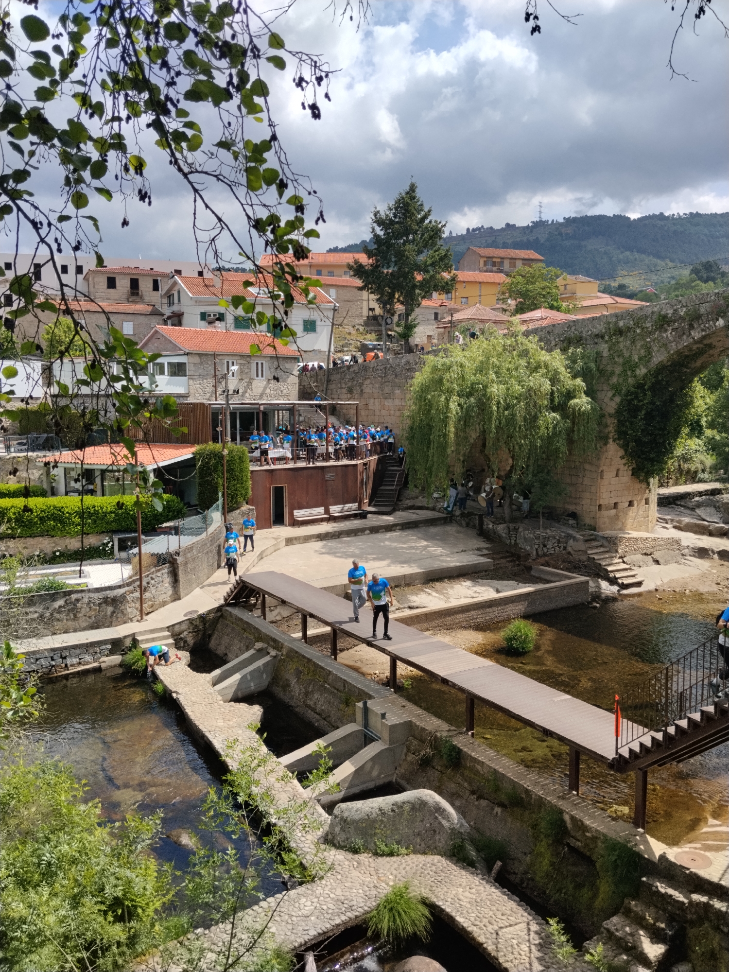The image shows a view of the village of Tarouca, Portugal, overlooking a stream and several bridges. In the background, there are buildings with orange tiled roofs. There is a group of people in blue shirts gathered on a terrace above the stream. A small group of musicians appear to be playing instruments near a stone bridge. People are crossing the stream using a wooden bridge. The sky is cloudy, and foliage frames the top and bottom of the view. The stream flows over a weir with concrete structures.