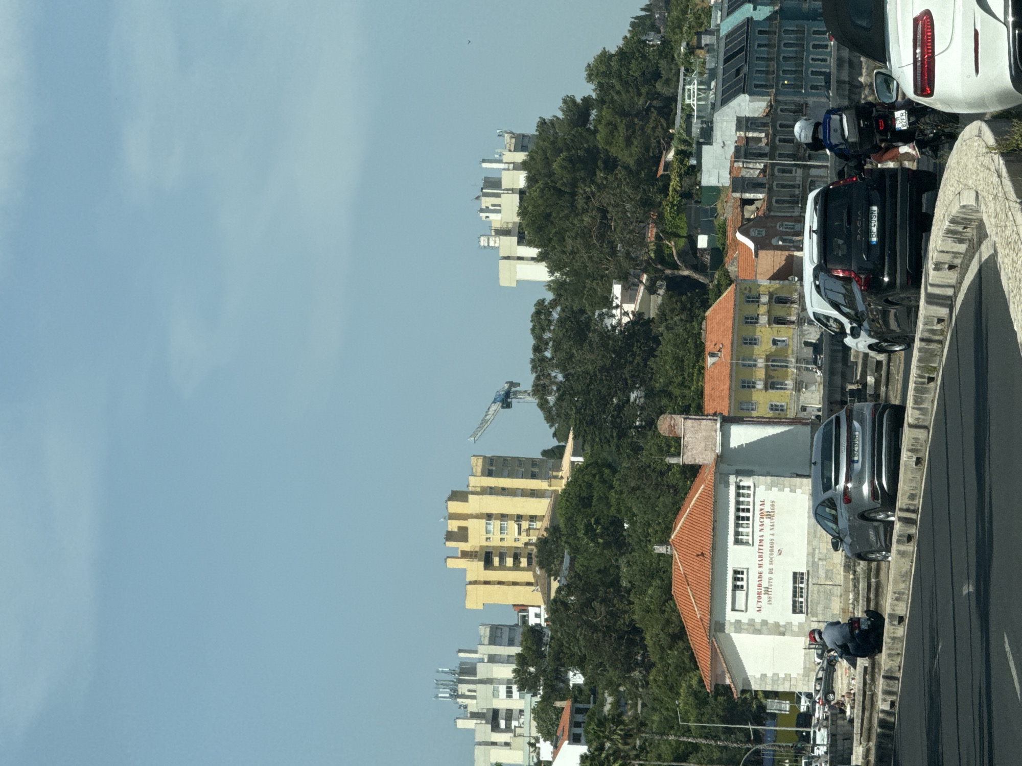 The image captures a scene on a sunny day in Oeiras, Portugal, likely along the Marginal road. In the foreground, a dark asphalt road curves to the left, with a concrete barrier separating it from a paved area. A silver Audi e-tron is parked on the paved area, facing left. To the left of the Audi, a person on a motorcycle wearing a blue shirt and red helmet is seen moving away from the camera. Further left, a few people are seated or lying on the ground near a white building with a red-tiled roof. This building has text that reads "AUTORIDADE MARÍTIMA NACIONAL" and "INSTITUTO DE SOCORROS A NÁUFRAGOS". Beyond this building, the landscape rises with lush green trees and several multi-story buildings, some with distinctive yellow and white facades. A construction crane is visible in the distance to the right. In the mid-ground and background, a mix of residential and commercial buildings are situated on a hillside, some exhibiting colorful facades. A white van and a black Dacia are also visible on the road. The sky is clear and blue.