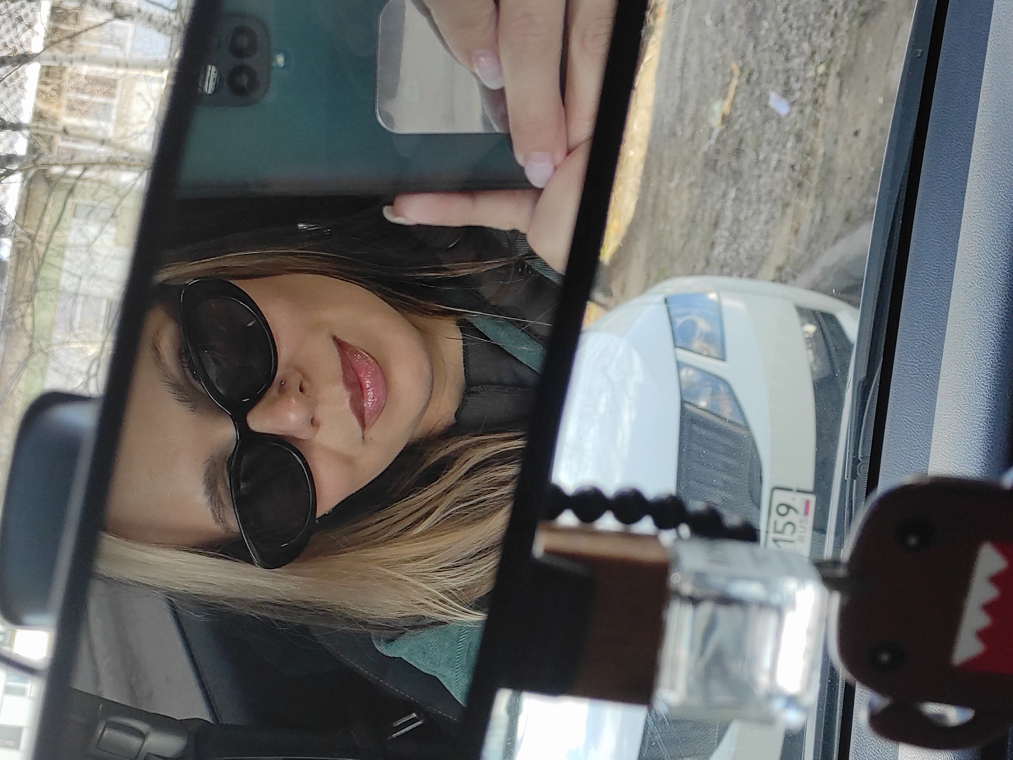 This vertical selfie, taken in a car's rearview mirror, captures a woman with blonde hair and sunglasses. Her face, partly obscured by the sunglasses, shows a nose piercing and glossy red lipstick, hinting at a slight smile. She is holding a smartphone with a teal/green case and a prominent camera array, reflecting her own image as she takes the photo. Her hands display light-colored nail polish.
To the right of the woman in the mirror's reflection, the front of a white car, possibly a Skoda, is visible. Its license plate clearly shows "159 RUS," identifying the vehicle as being from Russia's Perm Krai region, which aligns with the provided location of Chaikovsky. Outside the vehicle, the reflection reveals a street or road surface, along with blurred architectural elements and bare tree branches, indicating a daytime setting.
Hanging from the rearview mirror on the right side is a brown Domo-kun keychain. Alongside it, a string of black beads and a small, clear decorative item are also visible.