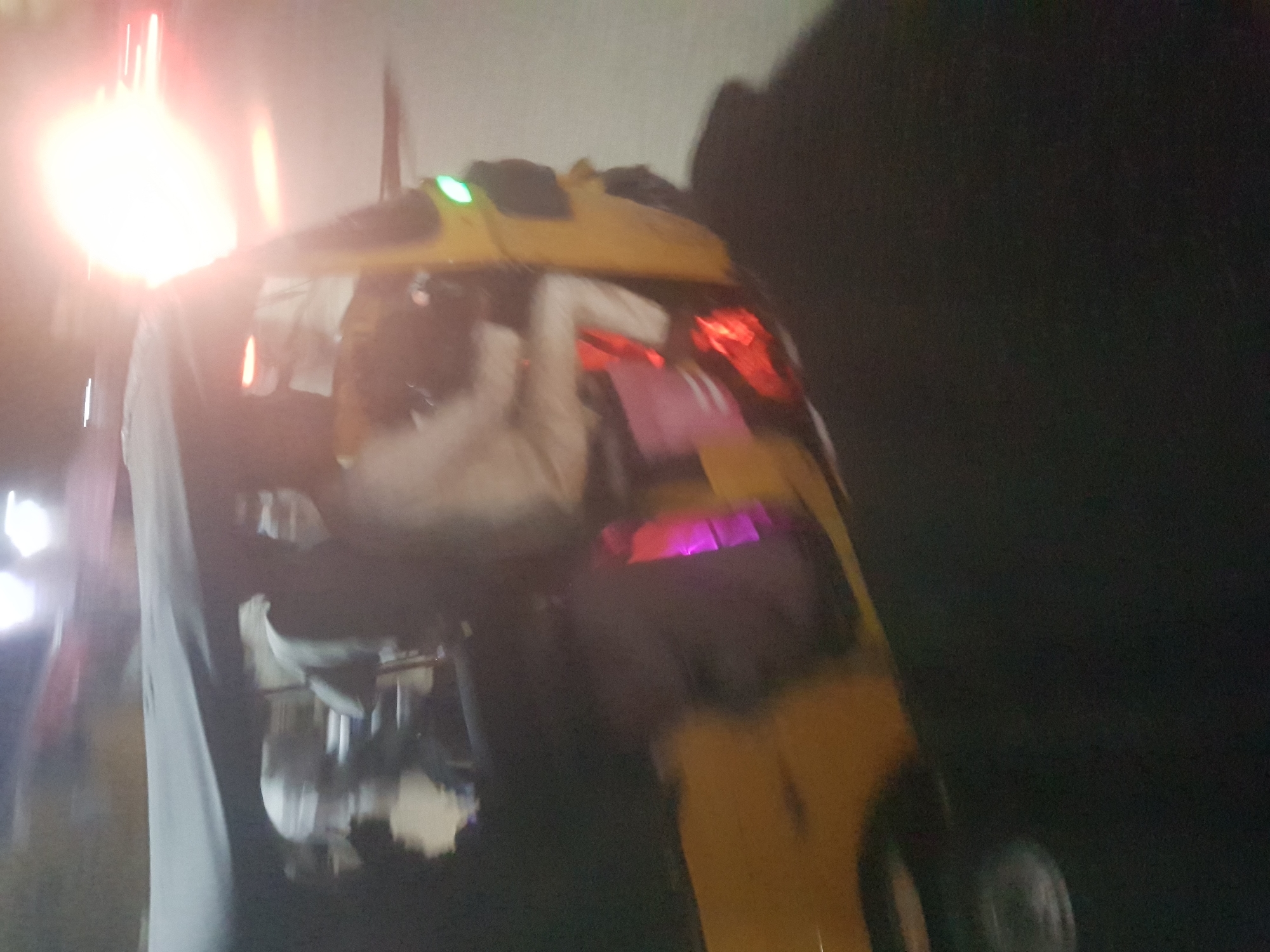 An overturned yellow vehicle, possibly an auto-rickshaw, is the central element. It is situated in an outdoor environment, likely at night, as suggested by the darkness. Light sources are visible, potentially streetlights and vehicle lights, causing significant lens flare. There appear to be people nearby, partially obscured and blurred due to motion or the lighting conditions. The location is possibly Dabawa, Nigeria.