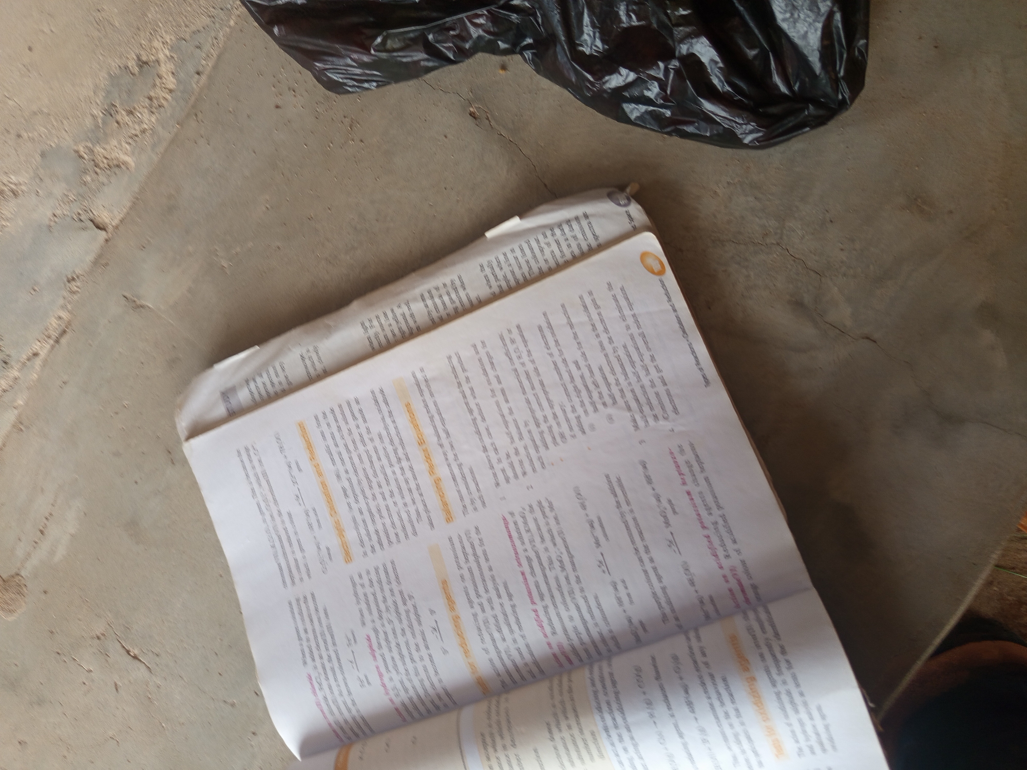 A close-up, slightly blurry, overhead shot focuses on an open book on a concrete floor. The book's pages are filled with text and chemical equations. To the right, a crumpled black plastic bag partially obscures the floor. The concrete floor is textured with cracks and speckled with sand or debris. The lighting is dim, creating shadows. The scene appears to be outdoors or in a sparsely furnished indoor space.