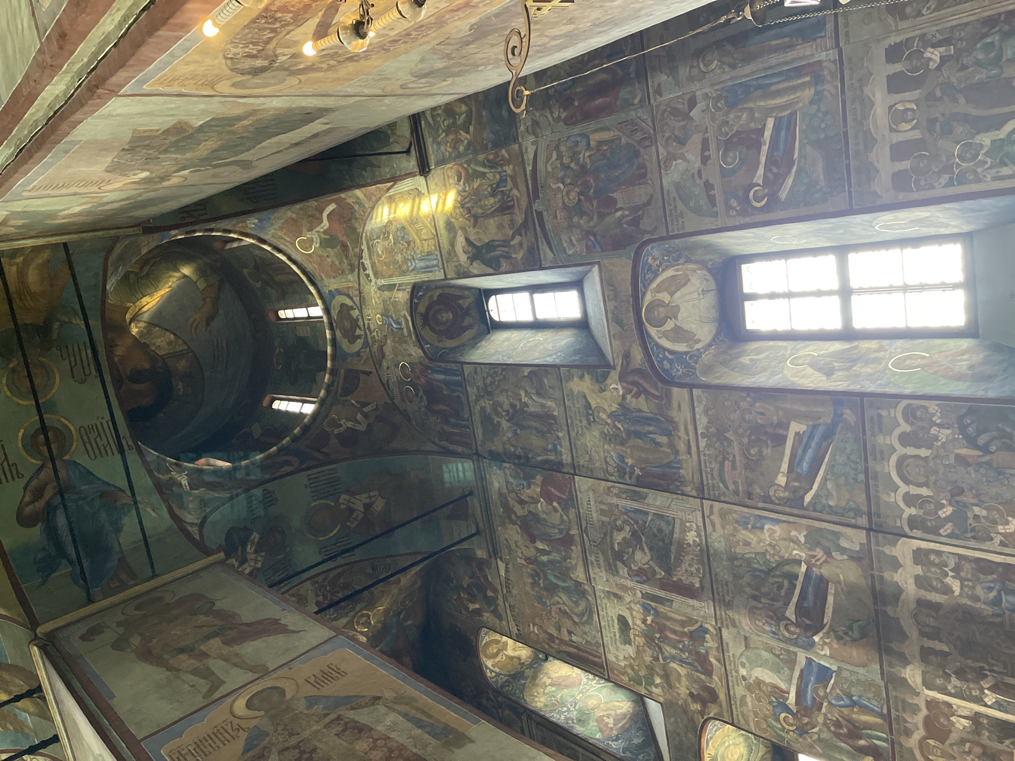 This is an interior photograph looking upwards into the dome and walls of a church, likely in Sergiyev Posad, Russia, given the style of the frescoes. The walls and ceiling are covered in intricate religious frescoes depicting biblical scenes and figures, rendered in a traditional Byzantine style with rich colors and gold accents. Sunlight streams through a tall, narrow window, illuminating portions of the frescoes and creating strong highlights and shadows. The architecture features soaring arches and a central dome. The frescoes are organized in registers, illustrating multiple narratives simultaneously. Some figures appear to be saints or angels, identifiable by halos. There are no visible people actively participating in the scene, but the frescoes themselves represent a multitude of human figures engaged in various narratives and contemplative poses. The overall impression is one of historical and spiritual grandeur.