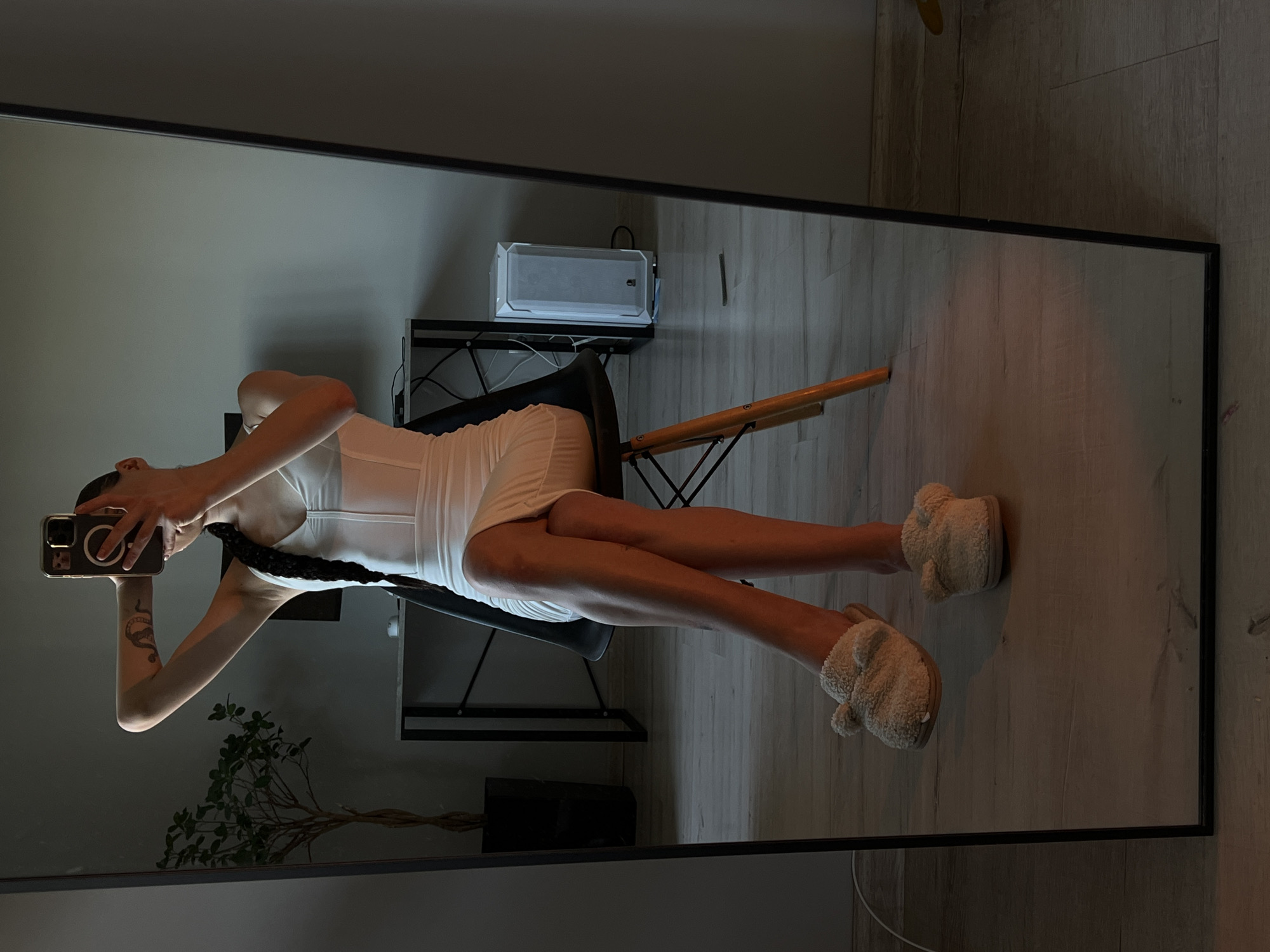 A woman is sitting on a chair and taking a selfie in a mirror in a room. She is wearing a white dress and fuzzy slippers. Her hair is in a braid. In the background, there is a desk with a computer, a plant, and a white computer tower. The floor is made of wood planks. The image is taken in Ternopil, Ukraine.