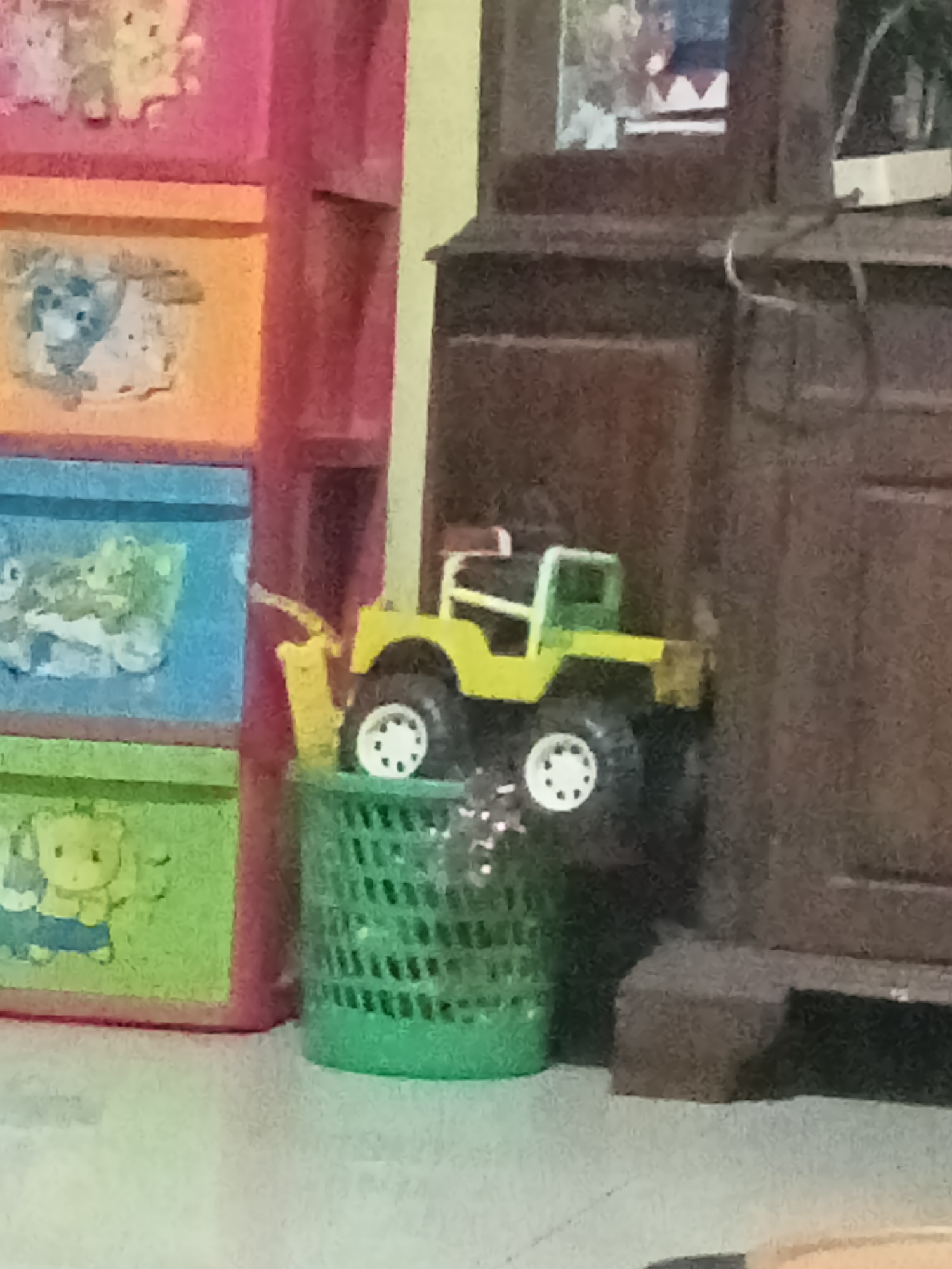 The image depicts a toy jeep, primarily yellow and green, placed inside a green plastic basket. The basket is situated next to a colorful toy storage unit with drawers of pink, orange, and blue, adorned with cartoon animal prints. To the right of the basket is a dark brown wooden furniture piece, possibly a cabinet or dresser, with some electronic wires visible on its surface. The scene appears to be indoors, likely in a domestic setting in Grabag, Indonesia. The lighting is somewhat dim, with a yellow cast. The overall composition suggests a casual arrangement of toys and furniture.
