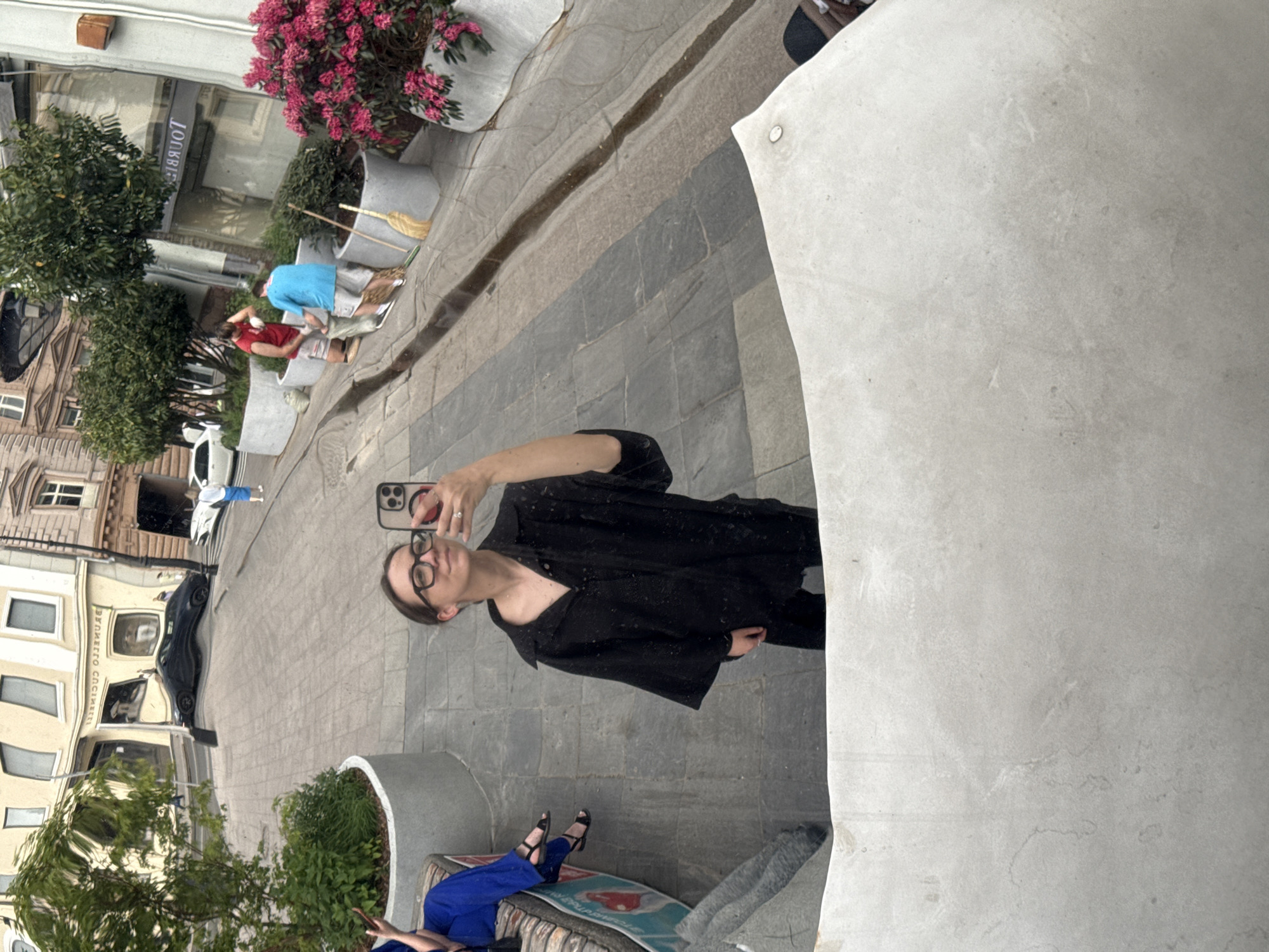 The image shows a mirrored reflection of a street scene, presumably in Moscow, Russia, based on the architectural styles and language visible.
In the foreground, a person with glasses is holding a smartphone, presumably taking a selfie, with their arm extended. They are wearing a black shirt. The reflection also shows a partial view of a person seated on a bench wearing blue pants and black sandals.
The reflection shows a street lined with buildings of mixed architectural styles, including classical-style buildings with multiple windows and ornate details. There's a row of planters with greenery, and pedestrians are visible in the background. There are also reflections of cars and storefronts. The word "BRUNETTO" is discernible on a building.
The overall composition of the image suggests the photographer is using a large, reflective surface, possibly a sculpture or architectural element, to capture the street scene and their own reflection.