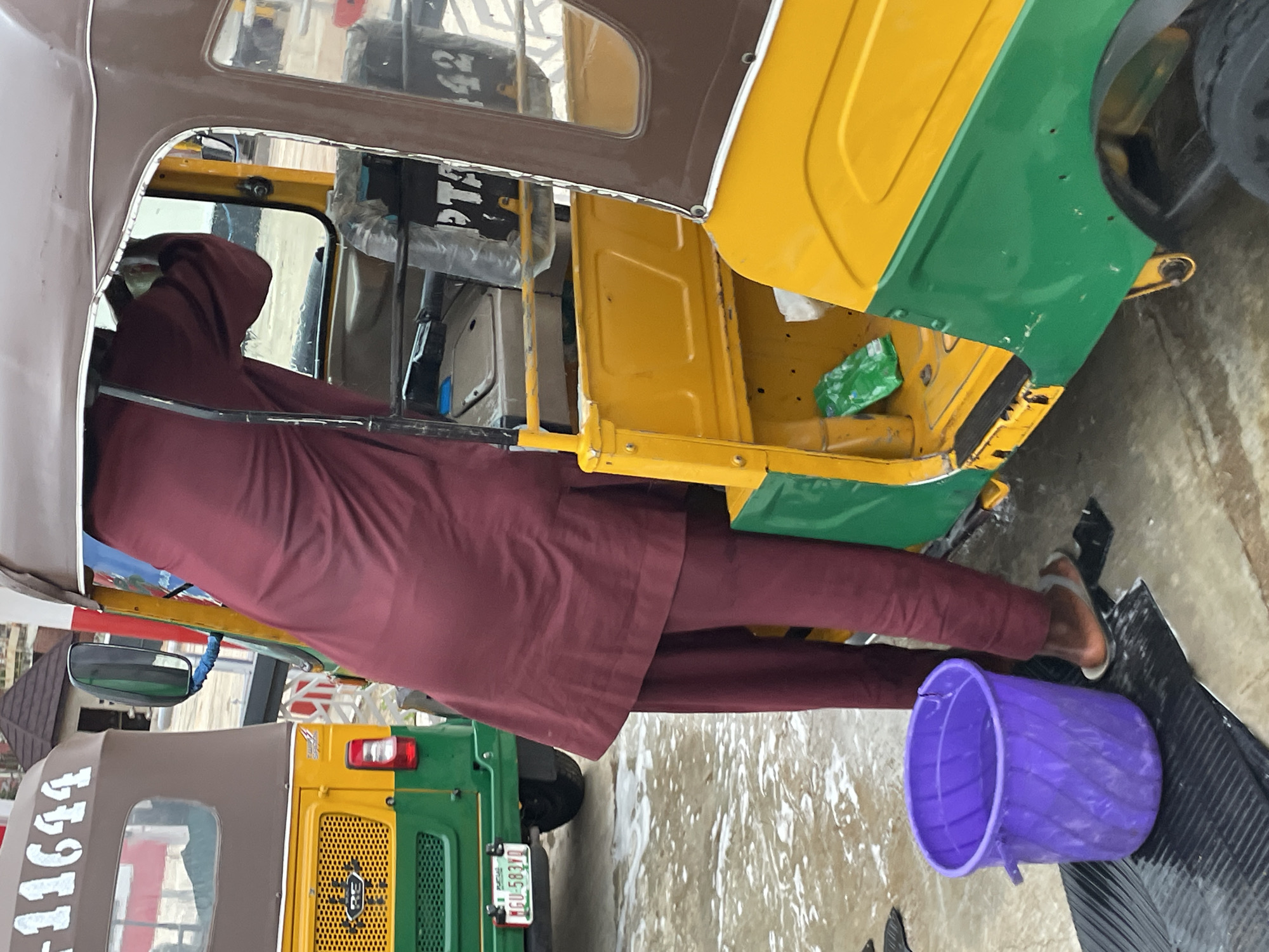 In Barakin Lerawi, Nigeria, a person wearing a maroon tunic and trousers stands next to a two-toned yellow and green auto-rickshaw. The auto-rickshaw has a brown fabric roof. The person is partially inside the vehicle, appearing to be attending to the interior. A purple bucket filled with water is placed on a black mat to the left of the auto-rickshaw. The ground is wet and appears to be a paved surface, possibly indicating recent washing or rain. Another auto-rickshaw, predominantly yellow and green, is visible in the background on the left. The scene suggests a moment of maintenance or cleaning of the auto-rickshaw.