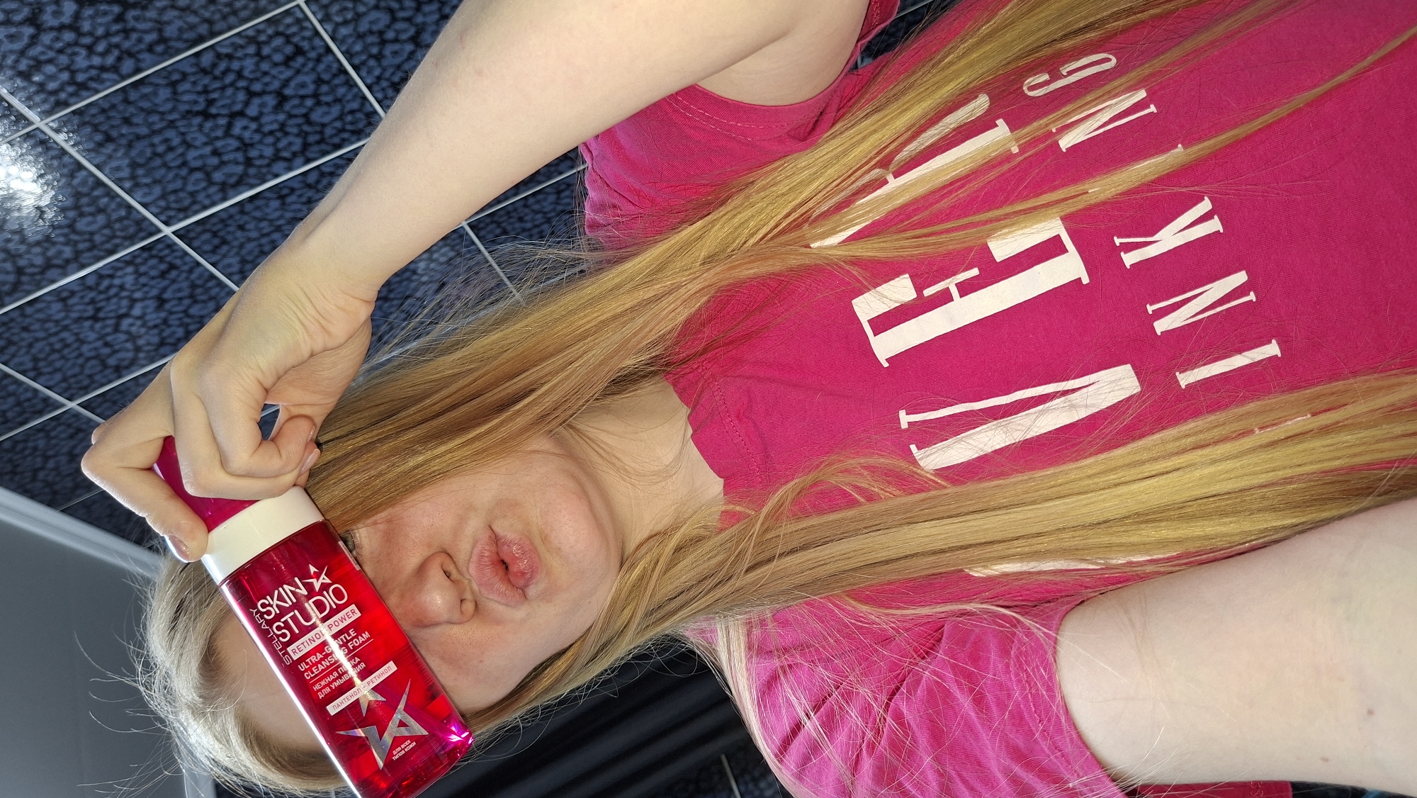A female holding a red and white bottle of Skin Studio retinol power ultra-gentle cleansing foam in front of her face is positioned diagonally against a background of black and blue patterned tiles. The woman, wearing a pink t-shirt with white text, has long blonde hair. The text on the bottle reads "Skin Studio Retinol Power Ultra-Gentle Cleansing Foam" in English and Russian. The location may be in the city of Chaikovsky, Russia.