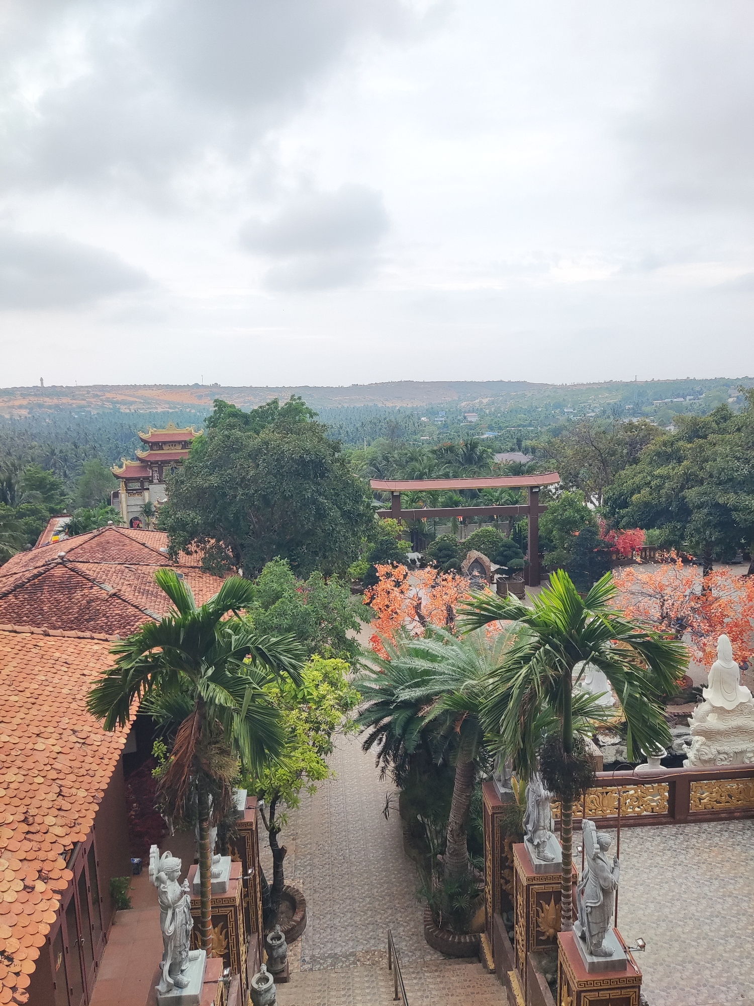 The image presents an elevated, panoramic view of a temple complex set within a vibrant, green landscape under an overcast sky. In the foreground left, a building with a distinctive red-tiled roof is partially visible. The mid-ground is dominated by lush tropical foliage, including various palm trees and large, verdant trees.
A prominent multi-tiered temple building with ornate golden-accented roofs stands centrally in the mid-ground, enveloped by greenery. To its right, a large, dark wooden torii-style gate opens to a meticulously landscaped garden. This garden features trees with striking bright orange-red foliage, smaller bonsai-like plants, and white animal statues. Further right, a large white Buddha statue sits peacefully in a paved courtyard, flanked by smaller stone figures along decorative, gold-patterned railings.
The background reveals undulating hills, some appearing sandy or arid, stretching towards the horizon. Beyond the temple grounds, a vast expanse of green trees, likely palm groves, extends across the landscape, with glimpses of distant structures.
The sky is a uniform, light grey, indicative of an overcast day with soft, diffused lighting. The absence of visible human activity contributes to a serene and contemplative ambiance. The architecture, statuary, and tropical environment strongly suggest a temple in East or Southeast Asia. Text is visible on a banner of the central temple building but is too indistinct to read.