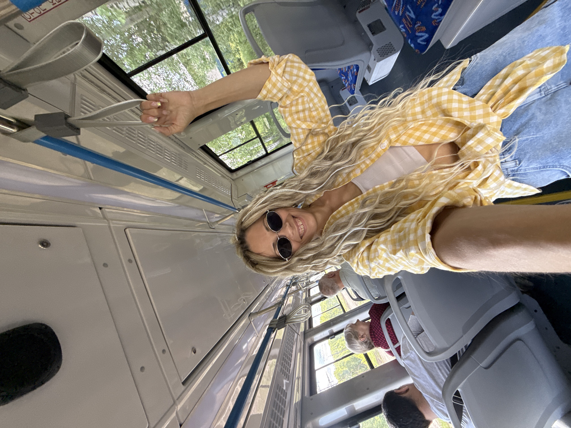 A woman with long blonde hair and round sunglasses is holding a grab handle in what appears to be a public bus. She is wearing a yellow and white plaid shirt over a white top, and denim jeans. The bus interior is mostly white and gray. Windows show trees outside. Two passengers are visible seated behind her. The location is Заволжье, Russia, as evidenced by Cyrillic text visible in a window reflection.