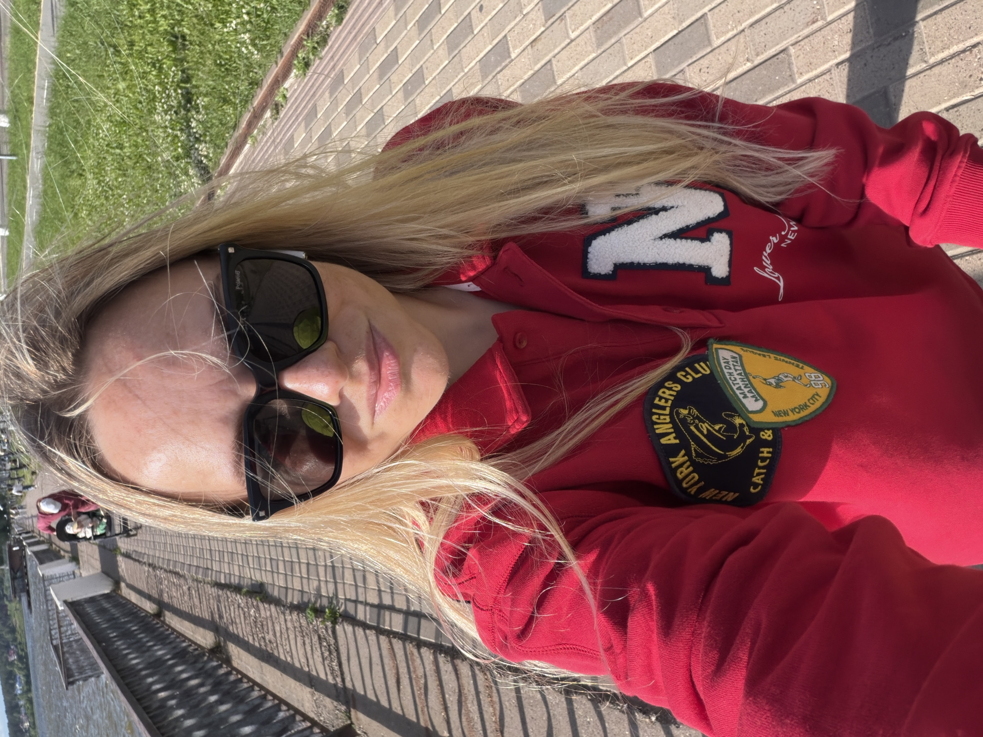 A blonde woman wearing sunglasses and a red sweatshirt with patches is taking a selfie outdoors. She is standing on a paved walkway next to a body of water, with a railing and trees to her left. In the background, a person is pushing a stroller with a child in it. The setting appears to be a park or promenade in the city of Votkinsk, Russia.
