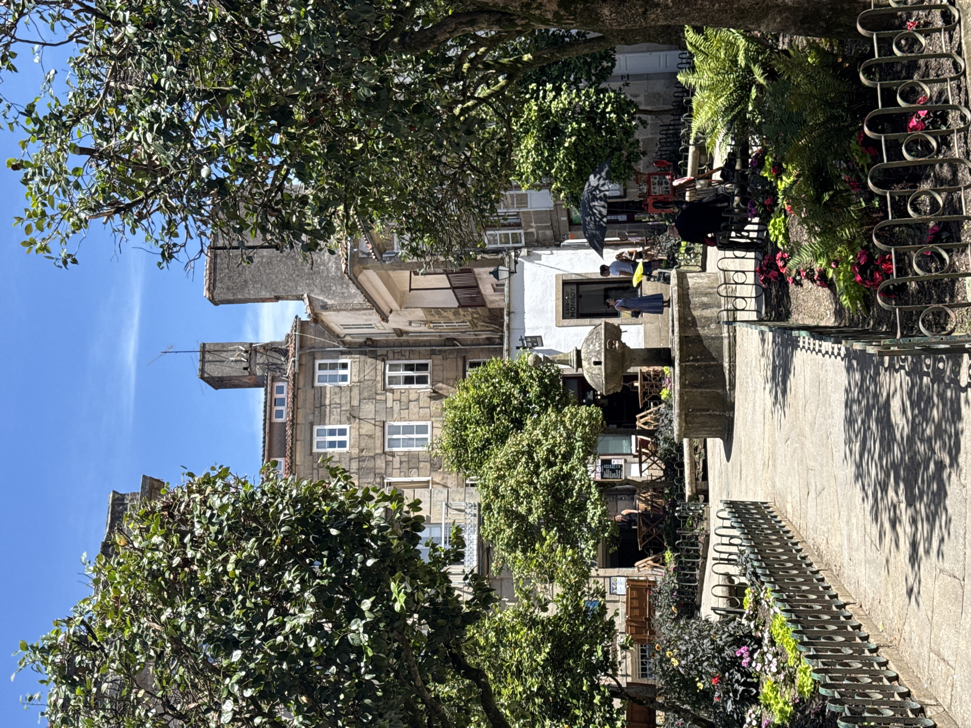 The image depicts a sunny day in a plaza in Santiago De Compostela, Spain, likely near the Mercado de Abastos or Praza de Cervantes, based on the architectural style of the surrounding buildings and the presence of outdoor seating for cafes. In the foreground, a paved pathway winds through a small park area with manicured flowerbeds bordered by decorative metal fencing. Lush green trees with full canopies flank the pathway, casting dappled shadows on the ground.
A stone fountain, the central feature of the plaza, stands in the mid-ground, surrounded by more trees and architectural elements. Behind the fountain, several buildings with stone facades and traditional windows are visible. Some of these appear to be cafes or shops, with outdoor tables and chairs set up. People are scattered throughout the scene, engaged in various activities. Two individuals stand near the fountain, while others are seated at tables or walking through the plaza. One person is seated on a bench in the park area, facing away from the camera. The sky is a clear, bright blue.
