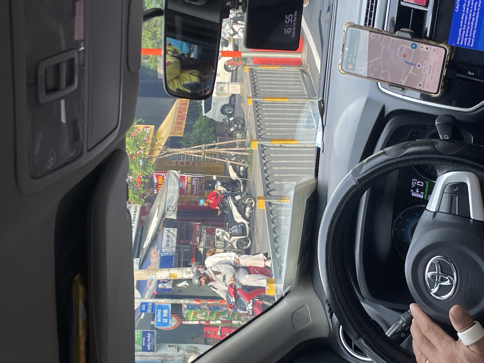 The image shows a street scene in Thủ Đức, Vietnam, as viewed from inside a car. In the foreground, a hand with a white ring on one finger is visible, resting on the steering wheel. The steering wheel has a Toyota logo. A smartphone mounted on the dashboard displays a navigation app with a map of the area and a route highlighted.
Outside the car, there is a busy street with various shops and buildings. Two people are riding a red scooter, wearing helmets. Several other scooters are parked or in motion. A white car and a white SUV are also visible on the street. Street signs in Vietnamese are present. The overall environment appears to be an urban setting during daylight. A dashcam is mounted on the rearview mirror, displaying "16:55" and "2023-05-19 Thu Sau".