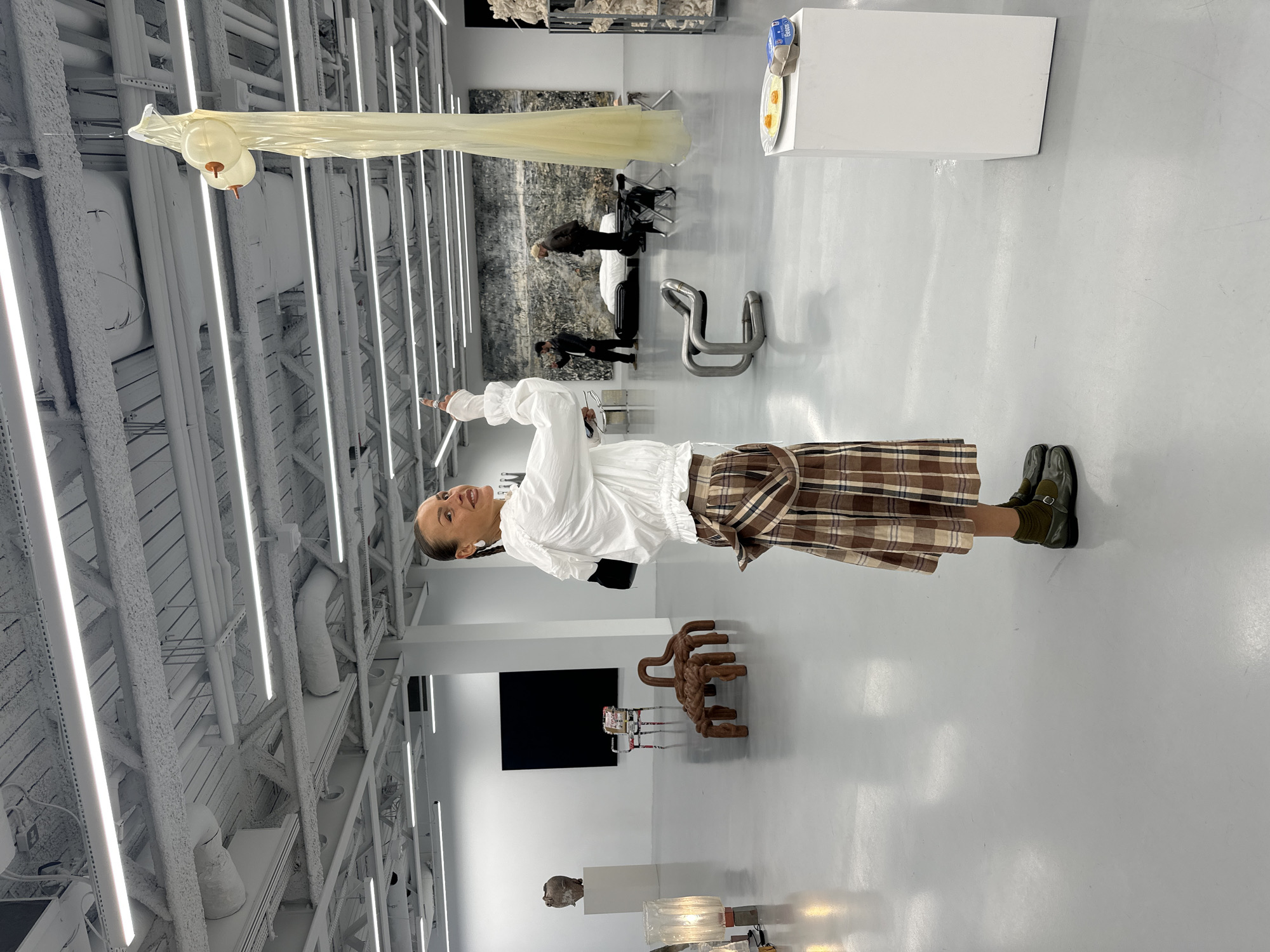 A woman in a white blouse and plaid skirt stands in an art gallery with a high ceiling featuring exposed ductwork, pipes, and lighting fixtures. She is pointing towards an installation that resembles a large, translucent object hanging from the ceiling. The gallery space includes several art pieces such as metal chairs, a black rectangle on the wall, and other abstract sculptures. People are walking in the background near a large artwork or mural. The setting suggests a modern or contemporary art exhibition. The city of London, United Kingdom, is not explicitly indicated.