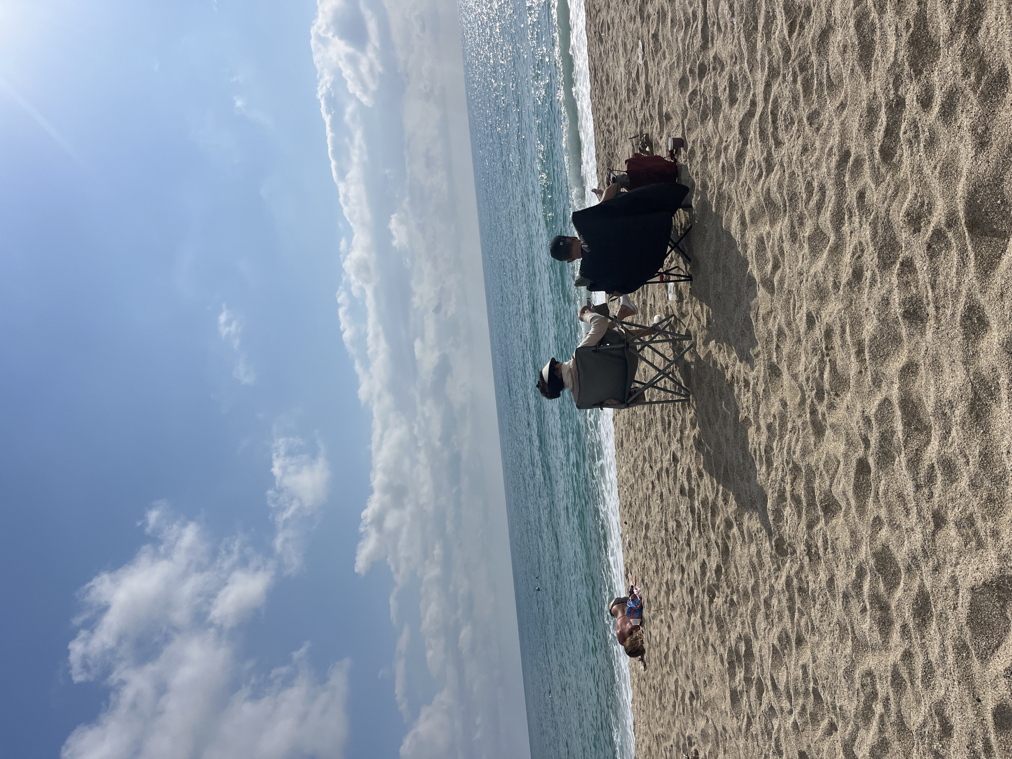 Two people are sitting in beach chairs on a sandy beach adjacent to the ocean under a partly cloudy sky. Another person is seen lying on the beach near the water. The location is likely Alanya, Turkey based on the geographical characteristics.