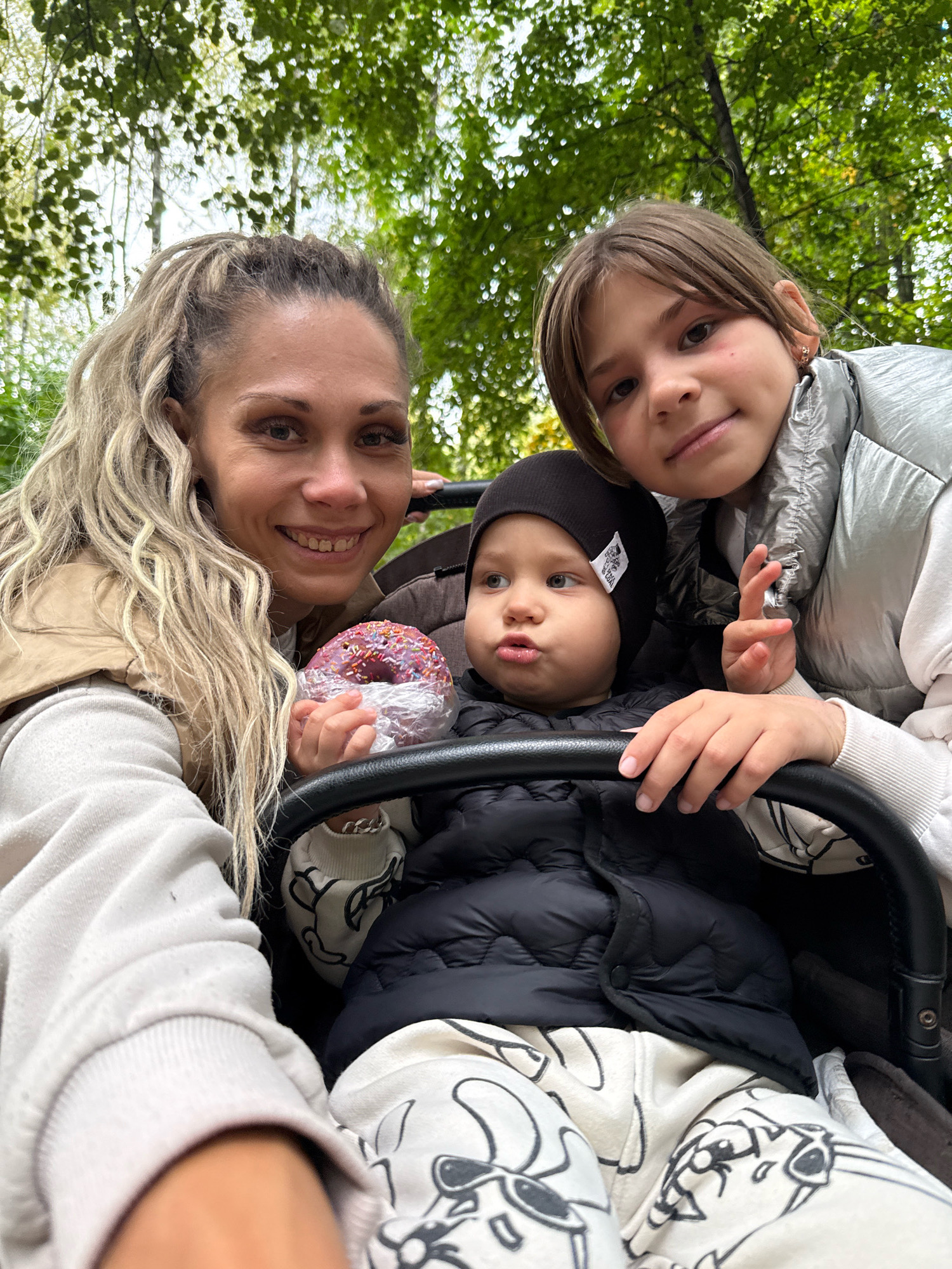 A selfie is taken outdoors in what appears to be a park or wooded area, with green trees and foliage in the background. Three people are visible. On the left, a woman with blonde hair pulled into a ponytail and wearing a tan vest over a cream sweatshirt smiles at the camera, holding a pink donut with sprinkles. In the center, a baby wearing a black beanie and a black puffer vest sits in a stroller. To the right, a young girl with brown hair, wearing a silver puffer vest, looks towards the camera. The stroller has a black frame. The image's timestamp and metadata are not provided, so a precise location within Rusanovo, Russia cannot be confirmed.