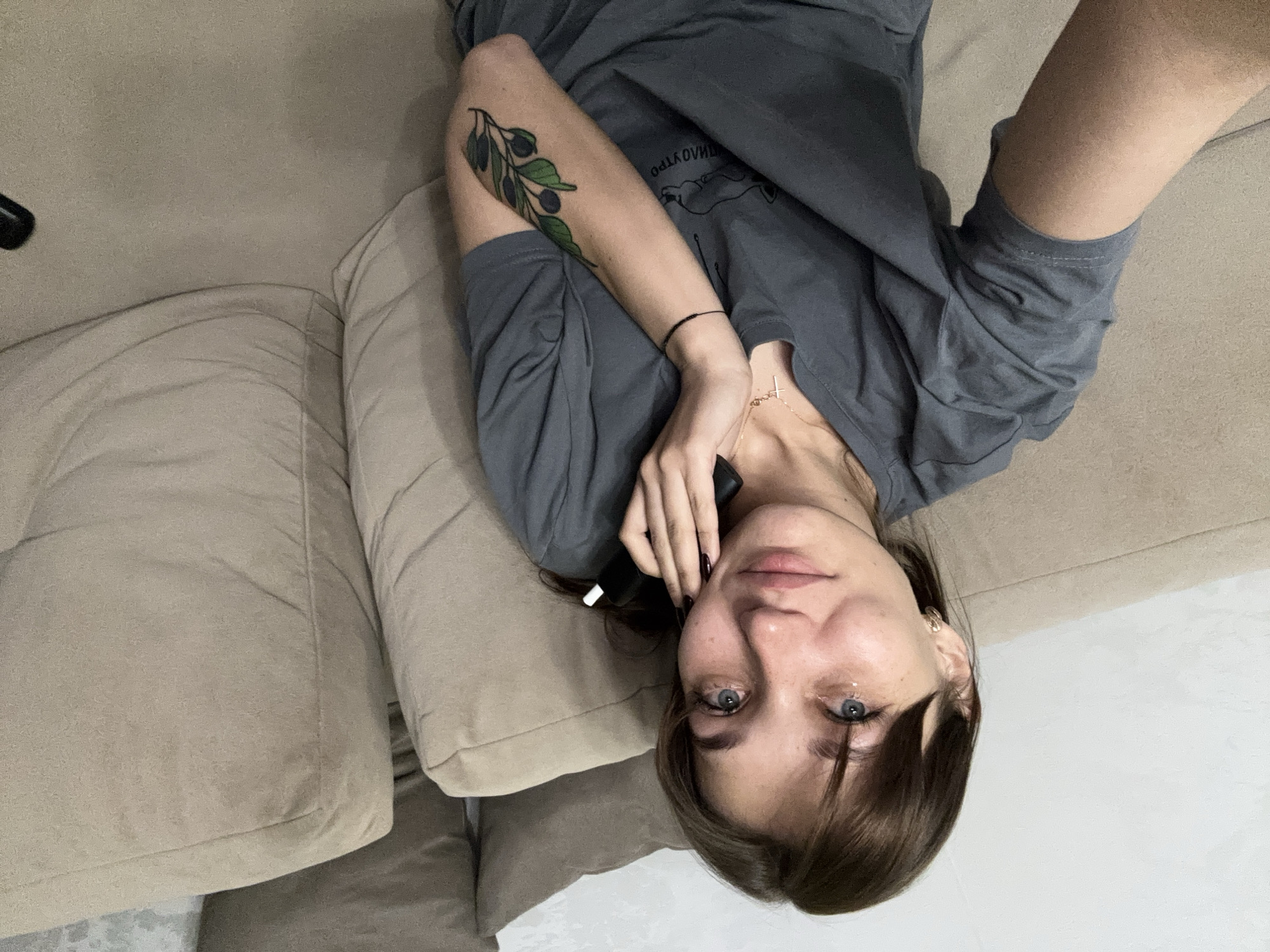 A young Caucasian woman with light brown hair and blue eyes is sitting on a beige sofa, holding a black object to her face with her right hand. She is wearing a grey t-shirt. She has a tattoo of a branch with leaves and berries on her left forearm. She is looking at the camera and appears to be crying, with a tear visible on her left cheek. The background consists of the sofa and a wall. The image was taken indoors in what appears to be a residential setting. There is no indication of the location being in Novokuznetsk, Russia.