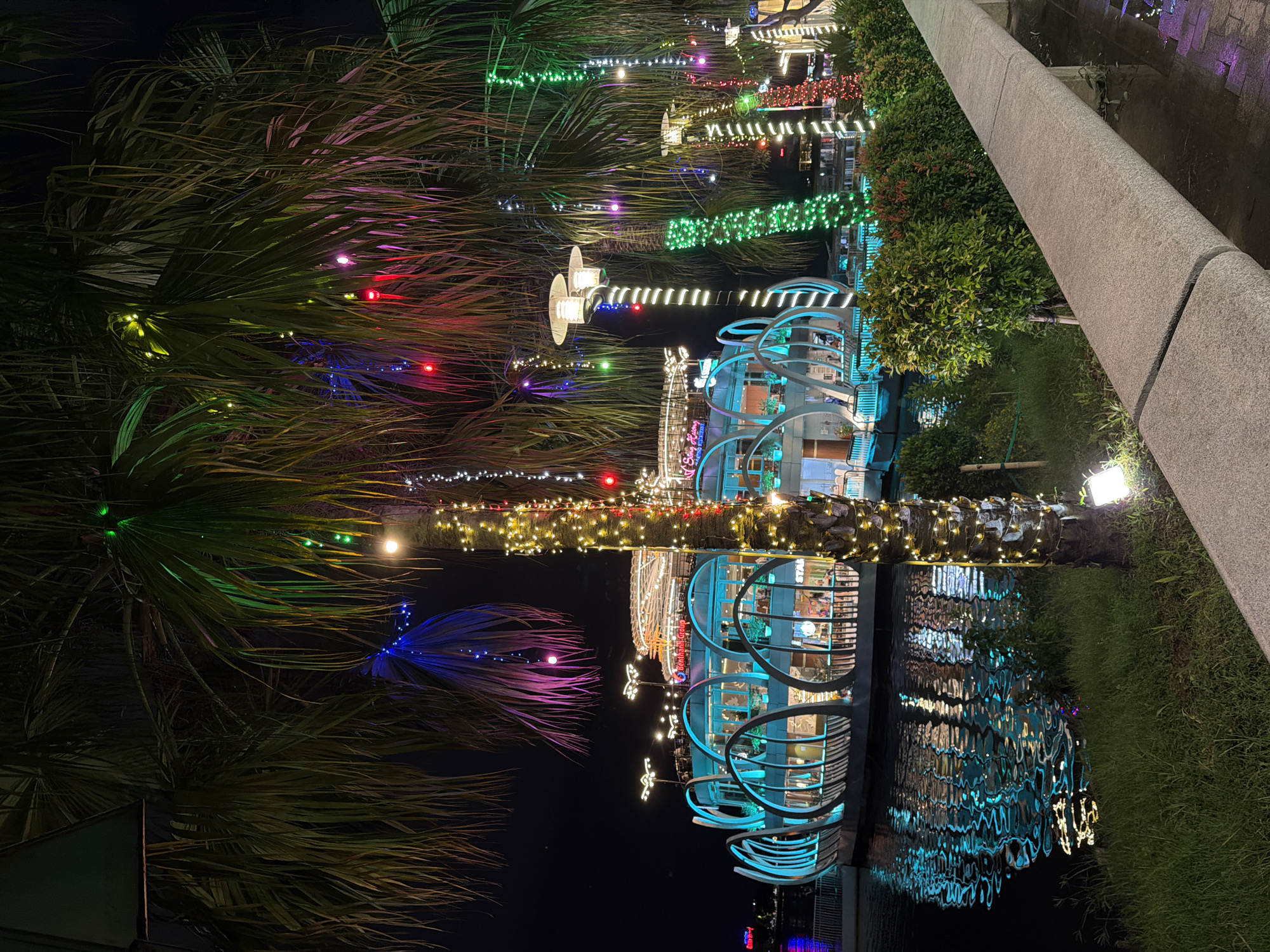 A brightly lit floating restaurant is situated on a body of water, reflecting its vibrant cyan and white lights onto the rippling surface. Palm trees adorned with strings of colorful LED lights, including green, blue, pink, and yellow, frame the scene, their fronds casting shadows against the dark night sky. The restaurant, with its distinctive curved architecture and multiple levels, appears to be a popular establishment. The foreground features a stone pathway and manicured green bushes, with grass bordering the water's edge. The overall ambiance suggests a festive or celebratory evening in a coastal or riverside urban setting within Thuận Hóa Ward, Vietnam, possibly a tourist destination or a lively entertainment district.