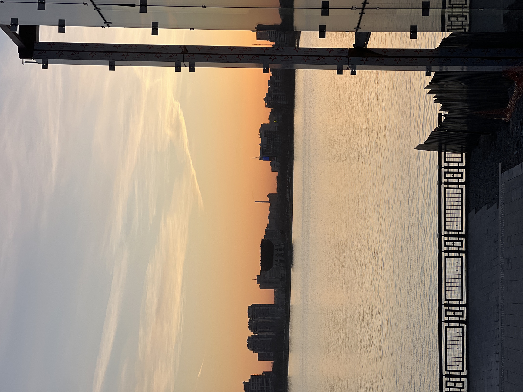 The image displays a serene urban waterfront scene, likely captured during either sunrise or sunset in Kazan, Russia. The upper portion of the frame is dominated by a sky exhibiting a beautiful gradient from a soft light blue at the top to a warm orange and yellow closer to the horizon. Wispy, elongated clouds are visible, illuminated by the low sun.
Running across the middle distance is the dark silhouette of a city skyline, featuring buildings of various heights against the colorful sky. Below the cityscape, a large body of water, possibly a river or lake, fills the lower part of the image, its surface gently rippled and reflecting the golden light of the sky.
In the immediate foreground, a modern architectural structure overhangs the scene from the top. It consists of glass panels supported by metal brackets and beams. On these metal beams, the Cyrillic text "АЛЮМИНСТРОЙ" (ALYUMINISTROY) is repeatedly visible alongside a star logo. Along the right edge of the water, a dark, ornate metal railing with geometric patterns defines the boundary of an embankment or promenade, next to a dark, paved area. The scene is devoid of people or specific activities, conveying a sense of tranquil contemplation and the quiet beauty of the golden hour.