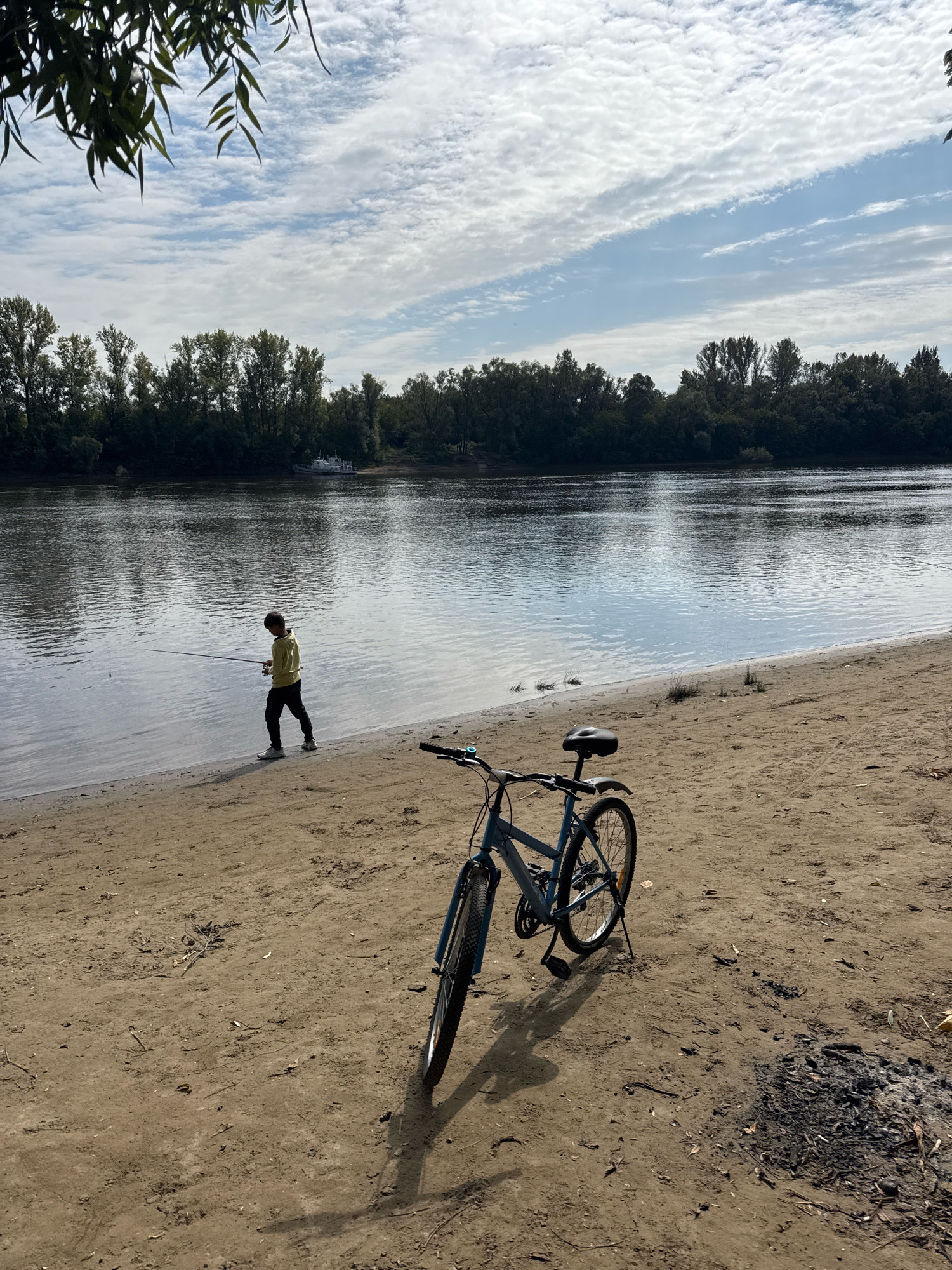 A boy is fishing on the bank of a wide, calm river. He is wearing a yellow long-sleeved shirt and black pants. A blue bicycle is parked on the sandy shore in the foreground, casting a shadow. In the background, across the river, is a dense line of green trees. The sky is partly cloudy with streaks of white clouds against a pale blue. The location is the Ufa river embankment in Ufa, Russia.