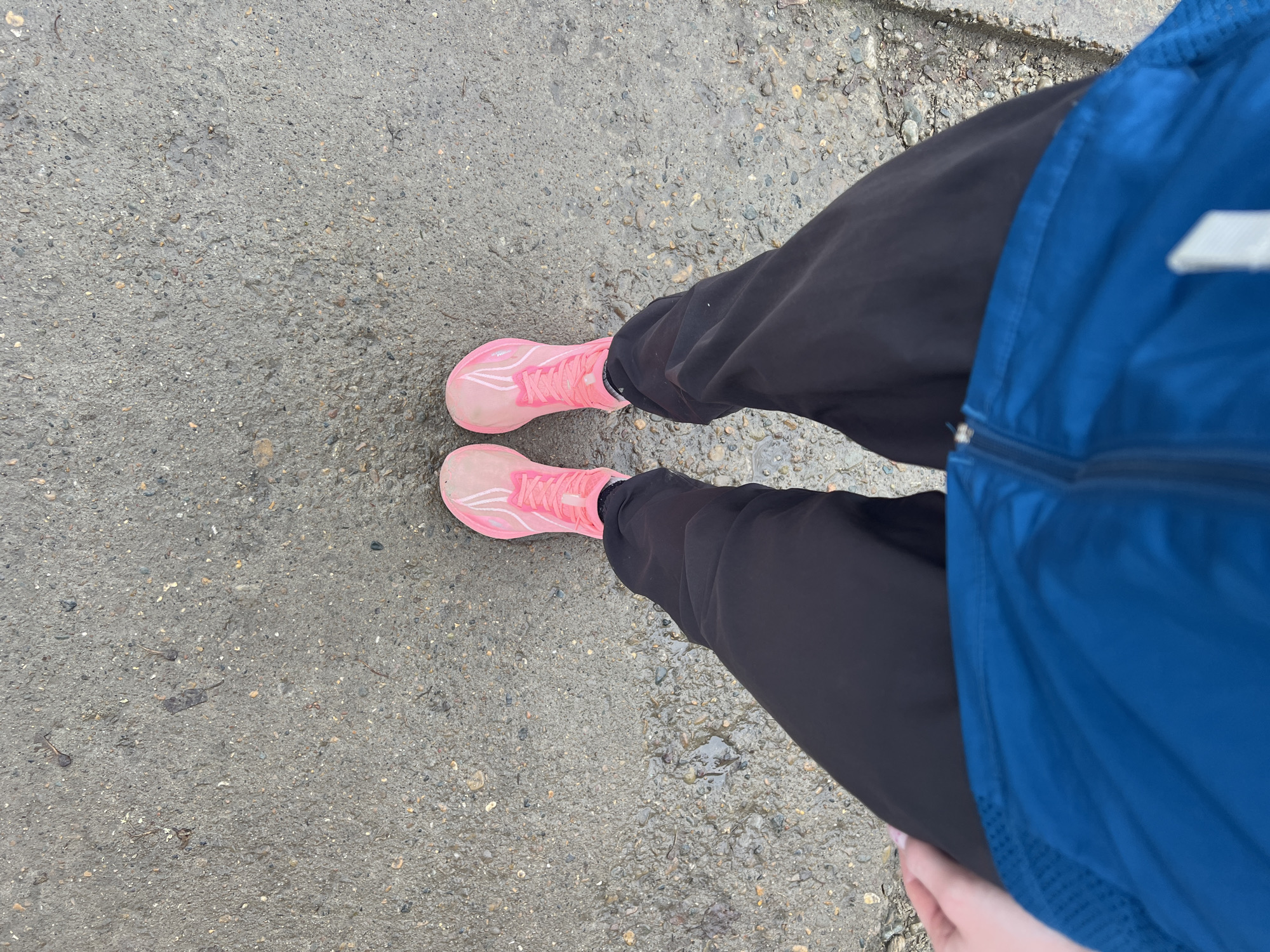 The image depicts a view from above of a person standing on a wet, paved surface. The person is wearing pink running shoes, black pants, and a blue jacket. The surface is light gray and appears to be asphalt or concrete with small pebbles embedded in it. The location is indeterminate without further information.